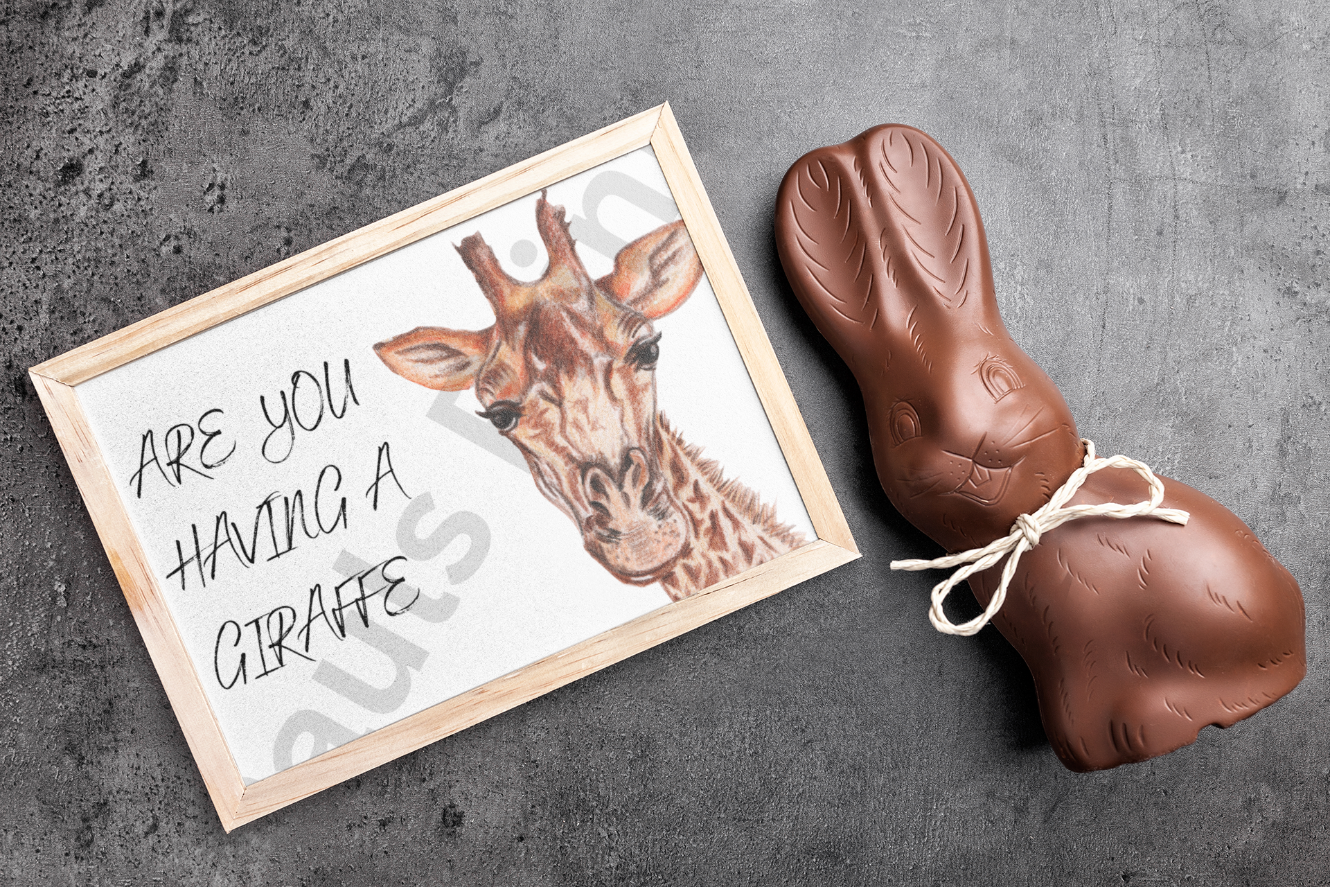 Humorous giraffe art print in a wooden frame placed next to a chocolate bunny on a dark stone surface.