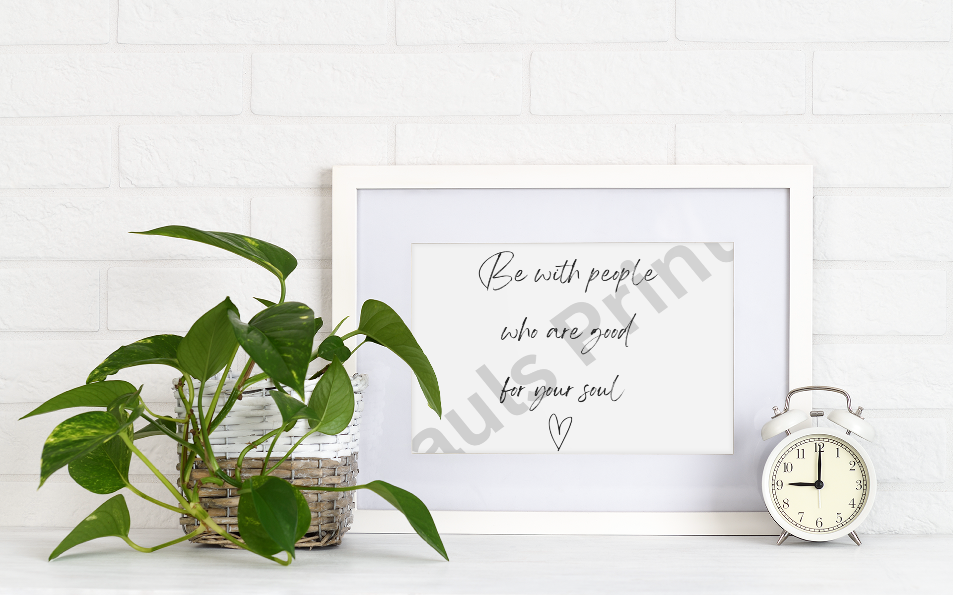 White frame holding a handwritten quote print beside a small clock and leafy houseplant on a white brick background