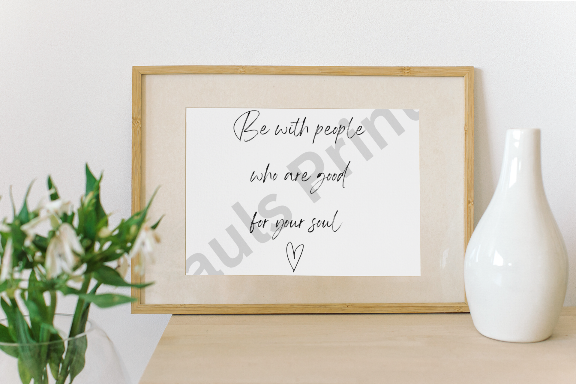 Wooden-framed quote print next to a ceramic white vase and flowers, on a light wood surface against a white wall