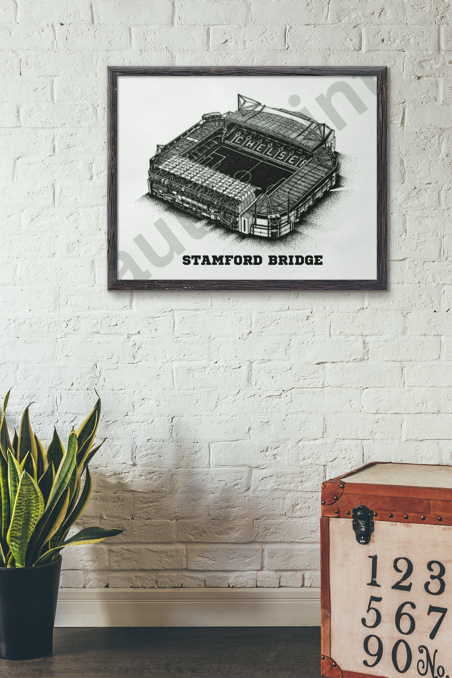 Framed black and white Stamford Bridge stadium print hanging on a white brick wall above a plant and vintage storage trunk.