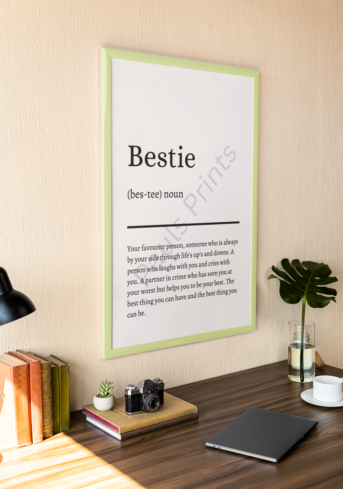 Light green-framed "Bestie" definition print hanging above a desk with books, a camera, a laptop, and a glass vase with a green monstera leaf.