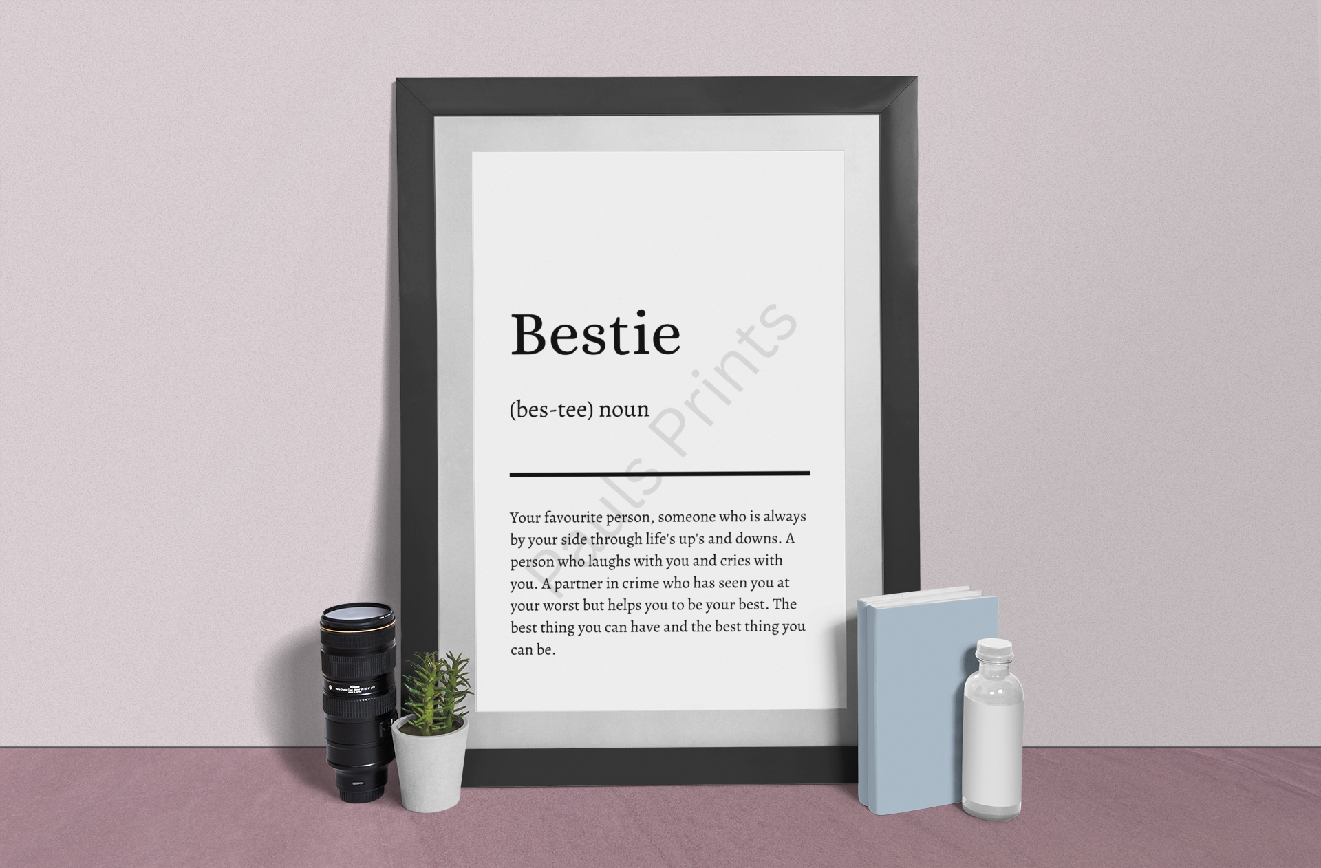 Black-framed "Bestie" definition print leaning against a pastel pink wall, styled with a camera lens, a succulent plant, and a blue book.