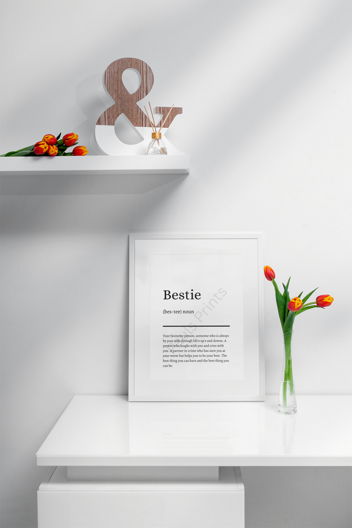 White-framed "Bestie" definition print on a sleek white cabinet, styled with a vase of orange tulips and a wooden ampersand decoration.