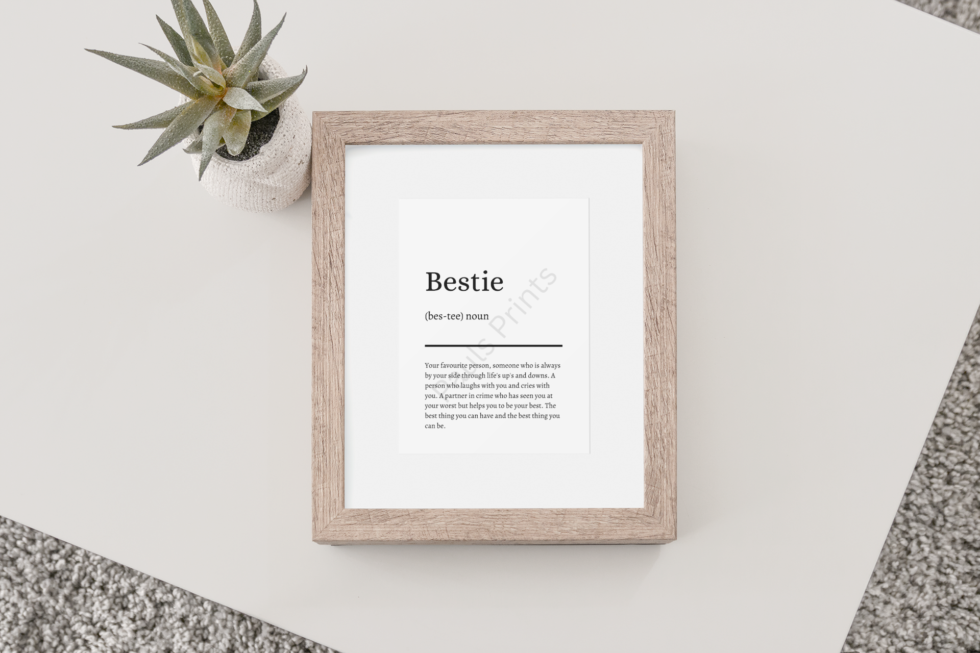 Wooden-framed "Bestie" definition print lying flat on a white surface beside a small succulent plant in a textured pot.