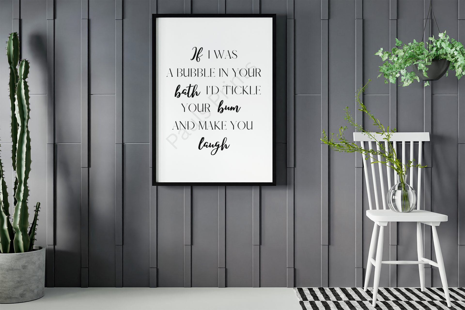 Black-framed humorous bathroom print with playful quote, hung on a dark grey wall next to a white chair with a vase and a tall cactus plant.