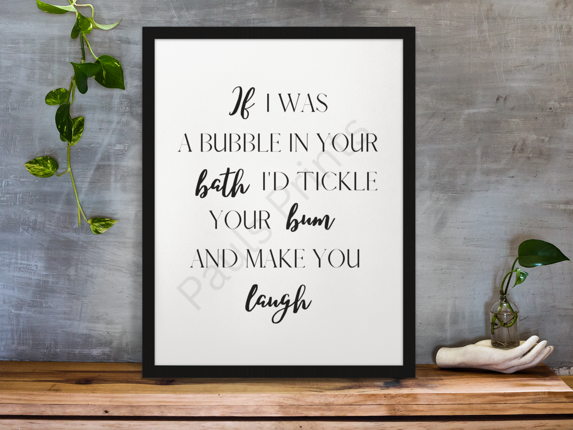 Black-framed humorous bathroom print with playful quote, displayed on a wooden surface against a grey wall with green vine plants.
