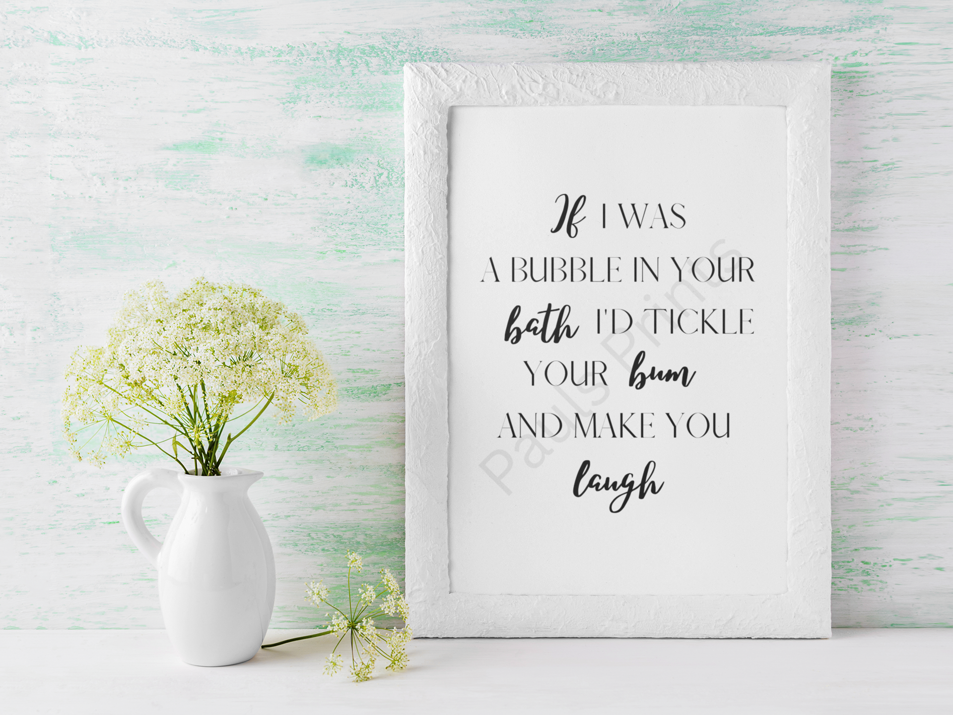 White-framed humorous bathroom print with playful quote, placed on a rustic whitewashed background next to a white jug of flowers.
