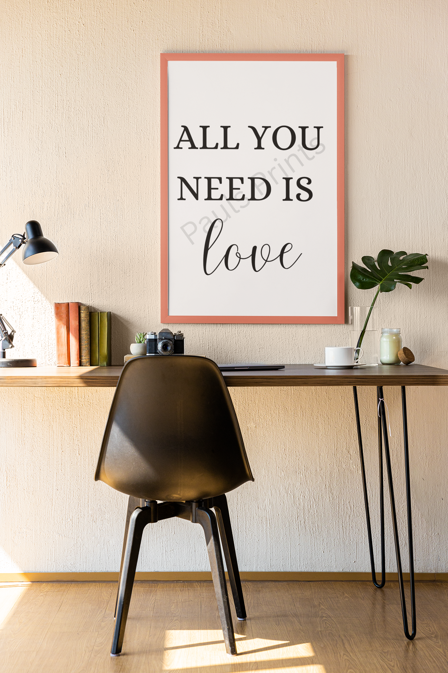 Coral-framed print with the words "All you need is love," hung above a desk with a black chair, books, a plant, and a coffee cup.