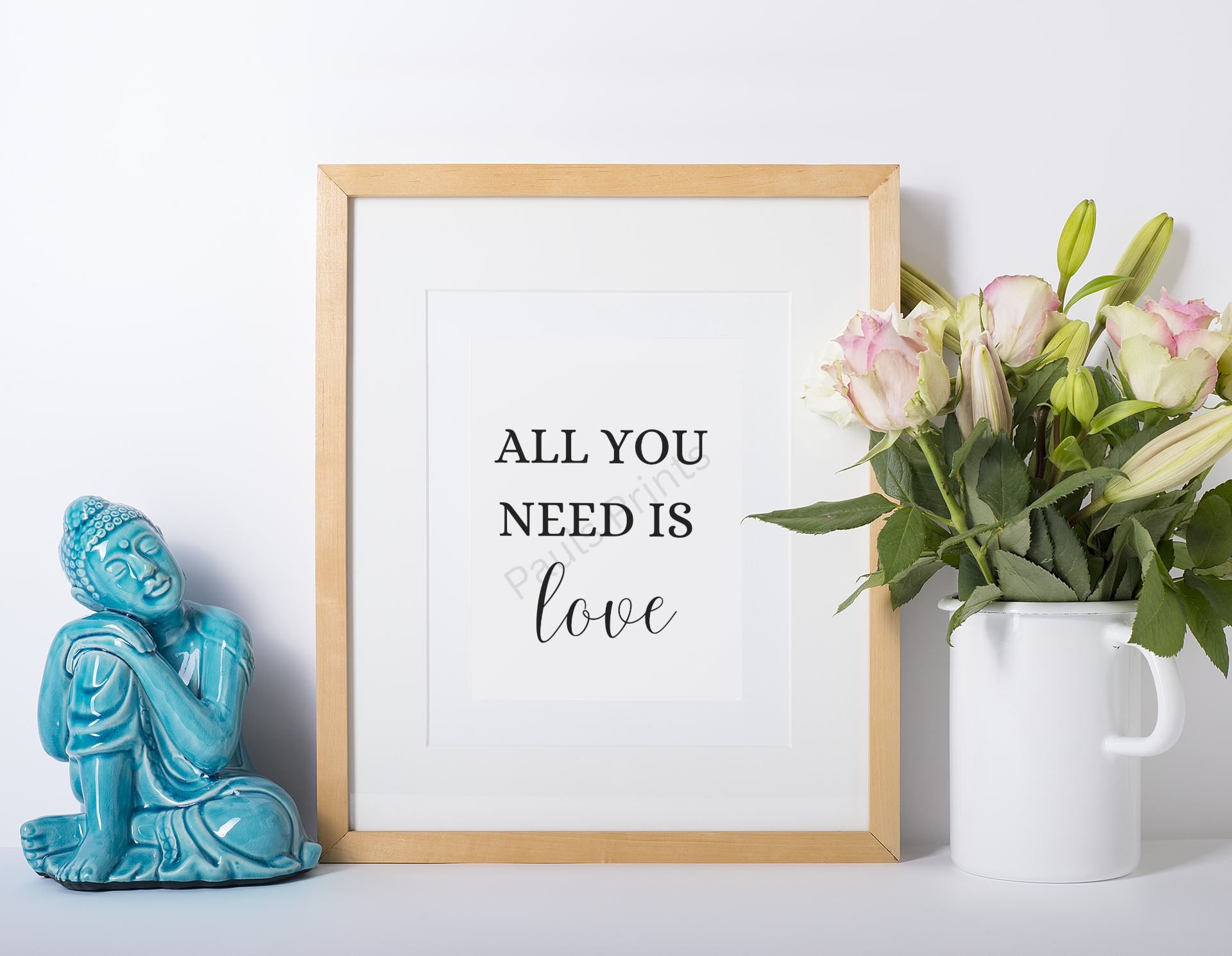 Wooden-framed print with the words "All you need is love," displayed between a turquoise Buddha ornament and a white jug of pink roses.