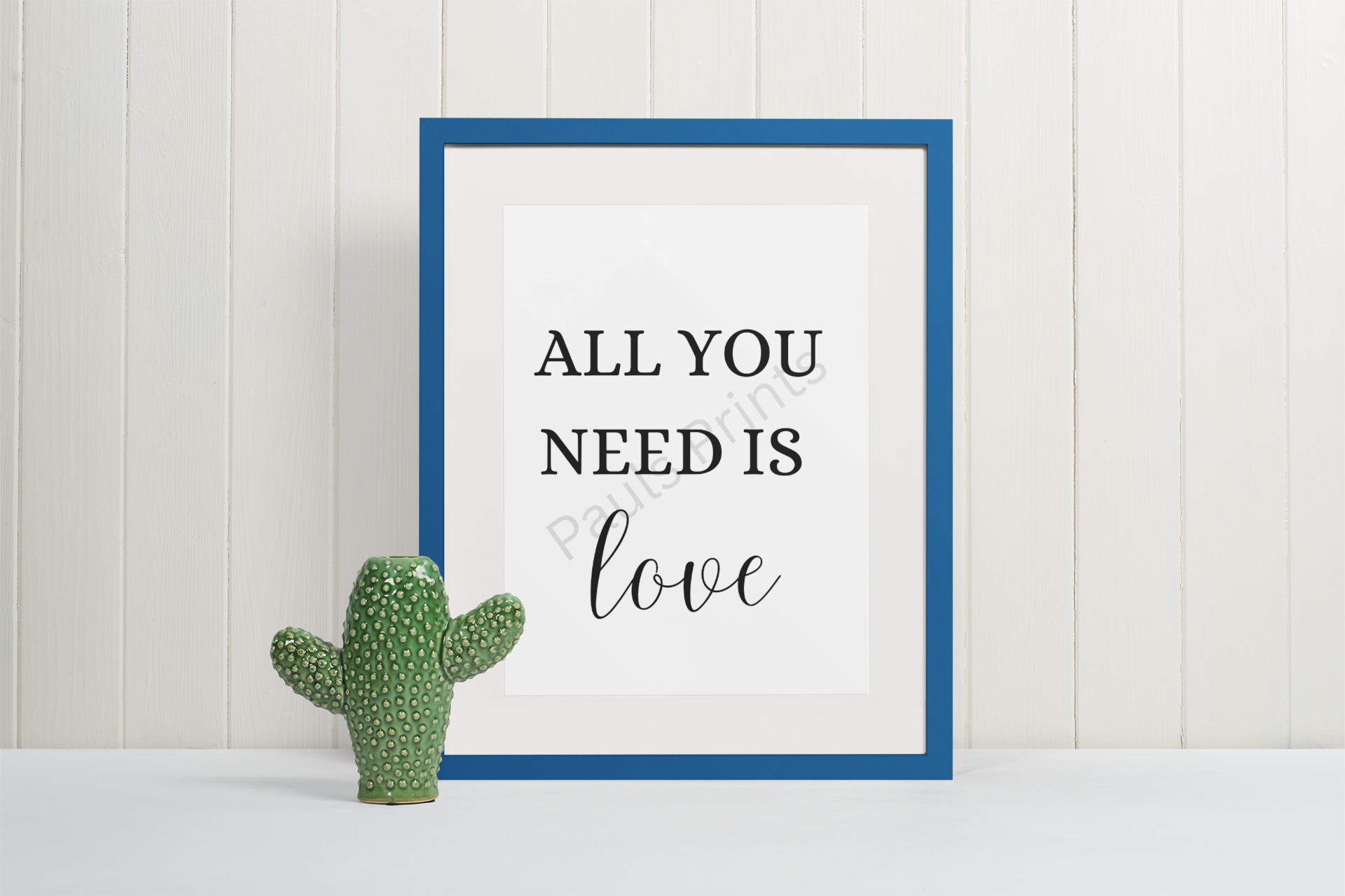 Blue-framed print with the words "All you need is love," standing against a white panelled wall beside a green cactus ornament.