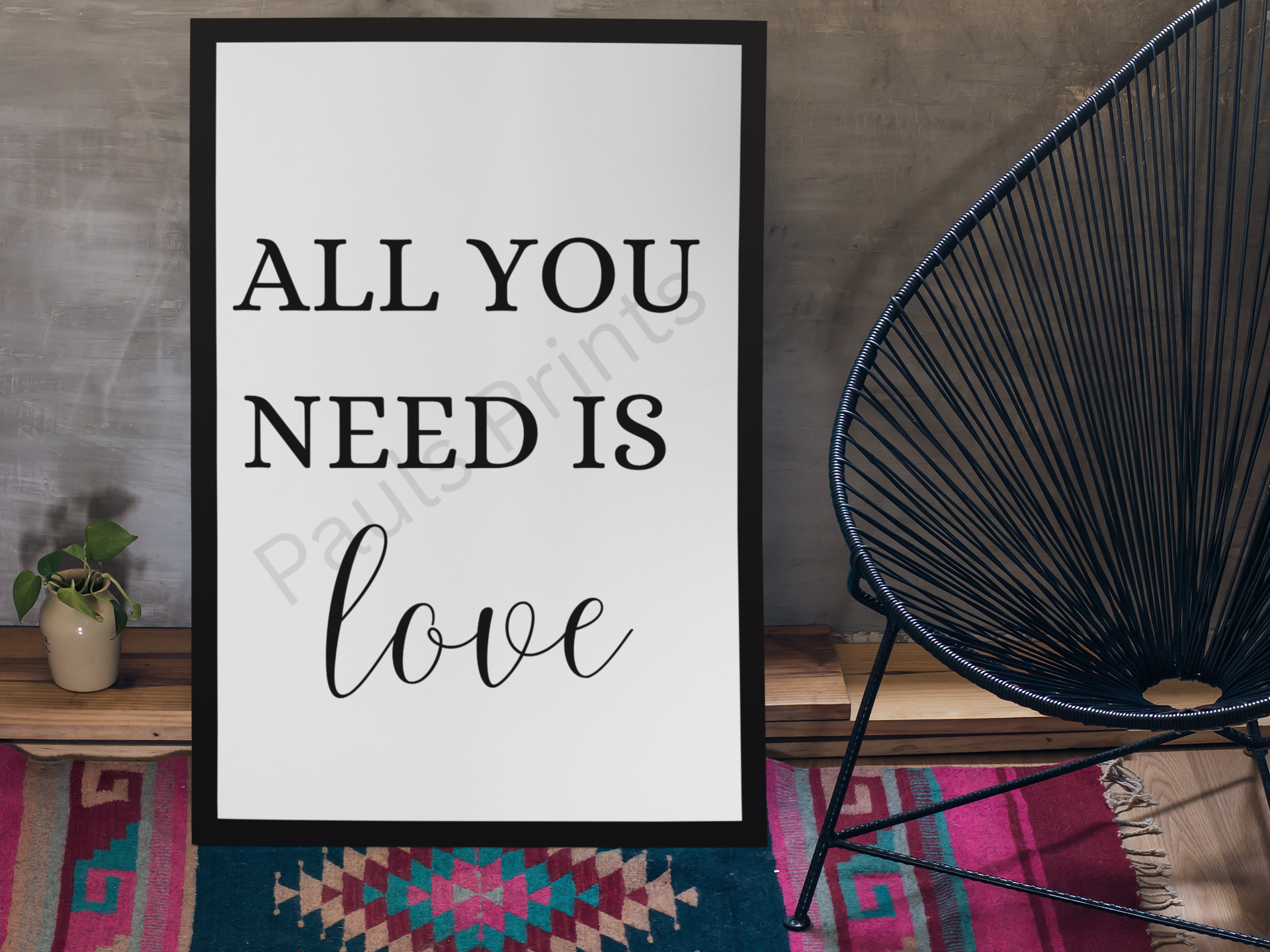 Black-framed print with the words "All you need is love" in bold and cursive fonts, leaning against a textured wall beside a black woven chair and a colourful patterned rug.