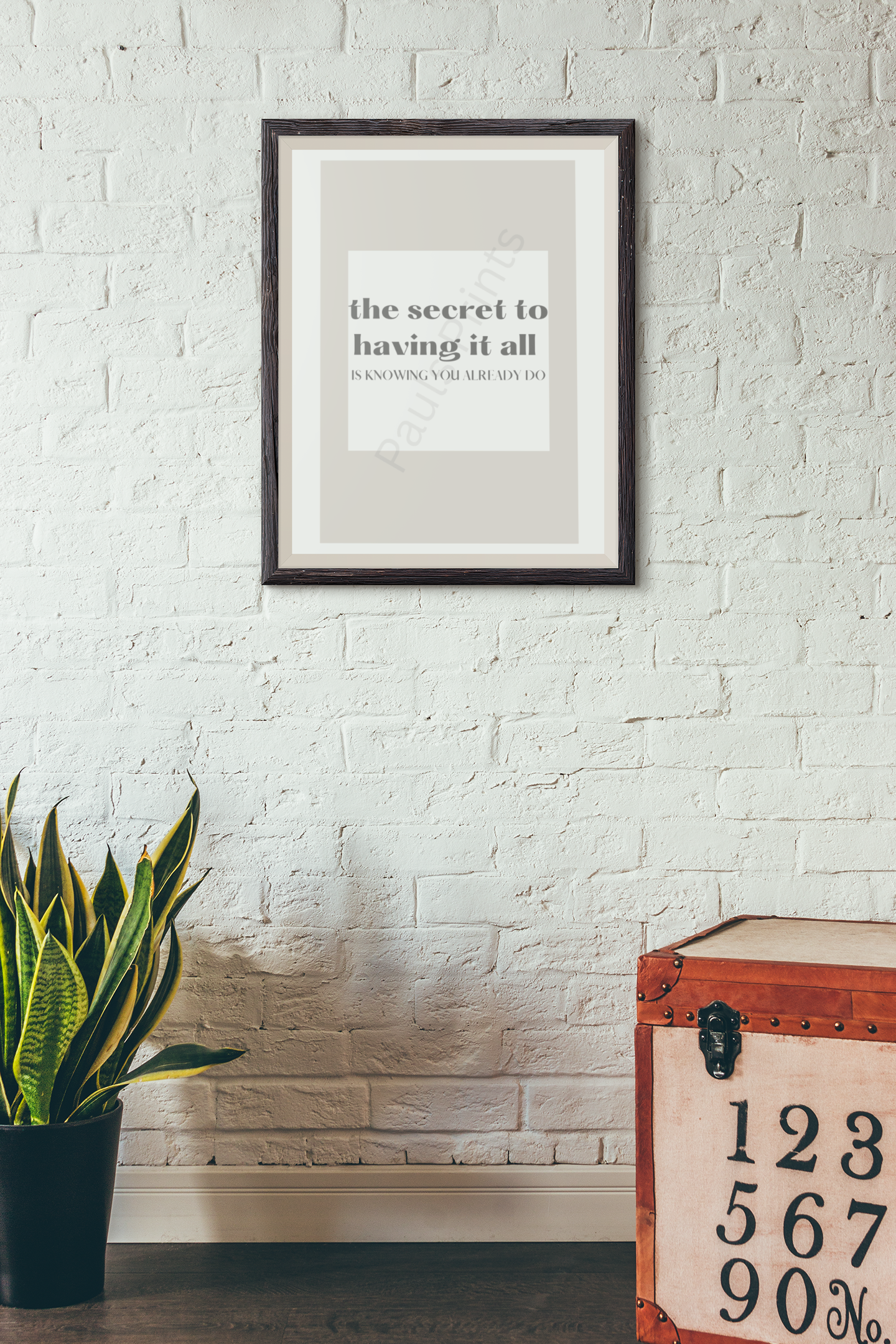 Dark wood-framed motivational print with the quote "The secret to having it all is knowing you already do," hung on a white brick wall beside a plant and a vintage-style chest.