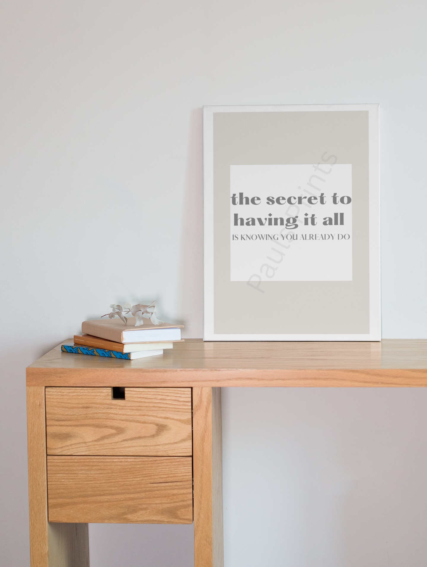 White-framed motivational print with the quote "The secret to having it all is knowing you already do," displayed on a wooden desk with stacked books and decorative items.