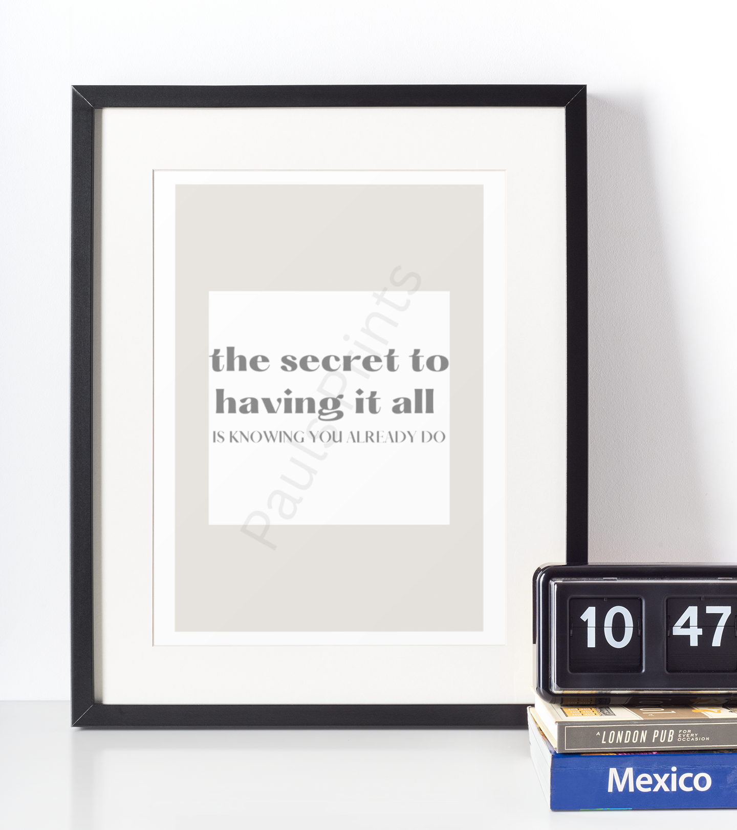 Black-framed motivational print with the quote "The secret to having it all is knowing you already do," placed beside a clock and books on a white surface.