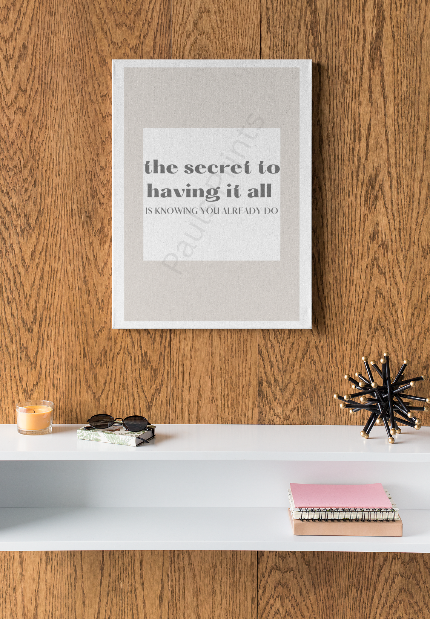 White-framed motivational print with the quote "The secret to having it all is knowing you already do," displayed on a wooden wall above a white shelf with candles, books, sunglasses, and decor.