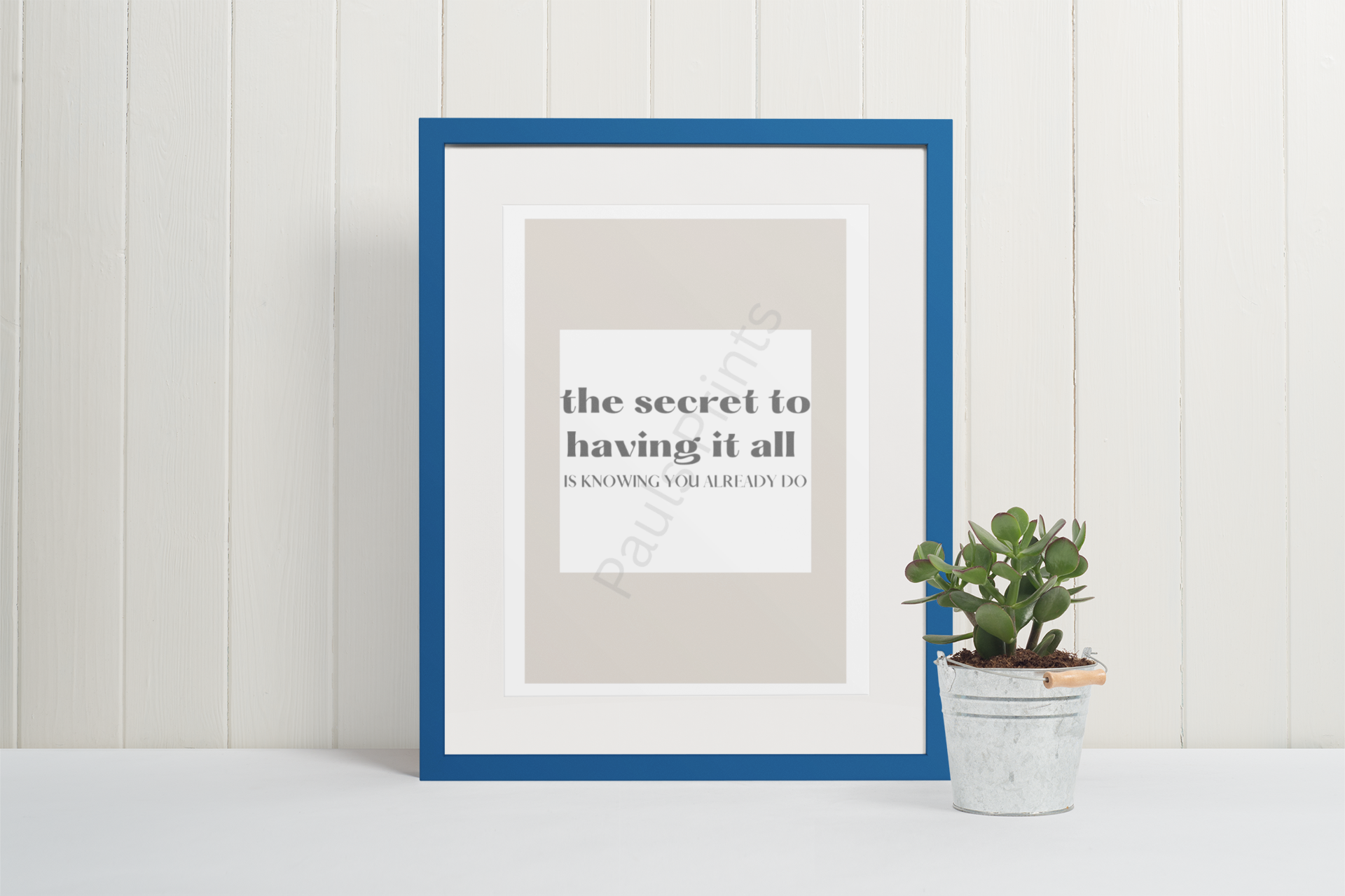 Blue-framed motivational print with the quote "The secret to having it all is knowing you already do," leaning against a white wall next to a potted succulent in a tin bucket.