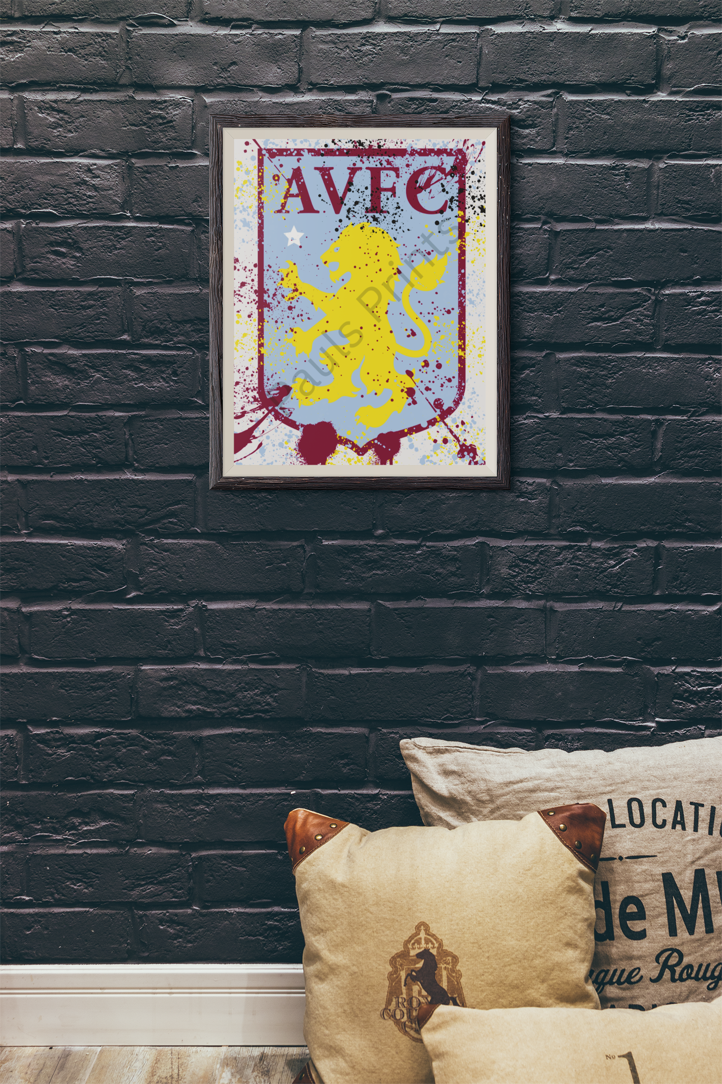 Framed Aston Villa FC crest artwork with yellow lion and claret lettering on a splatter background, hanging on a dark brick wall above cushions.