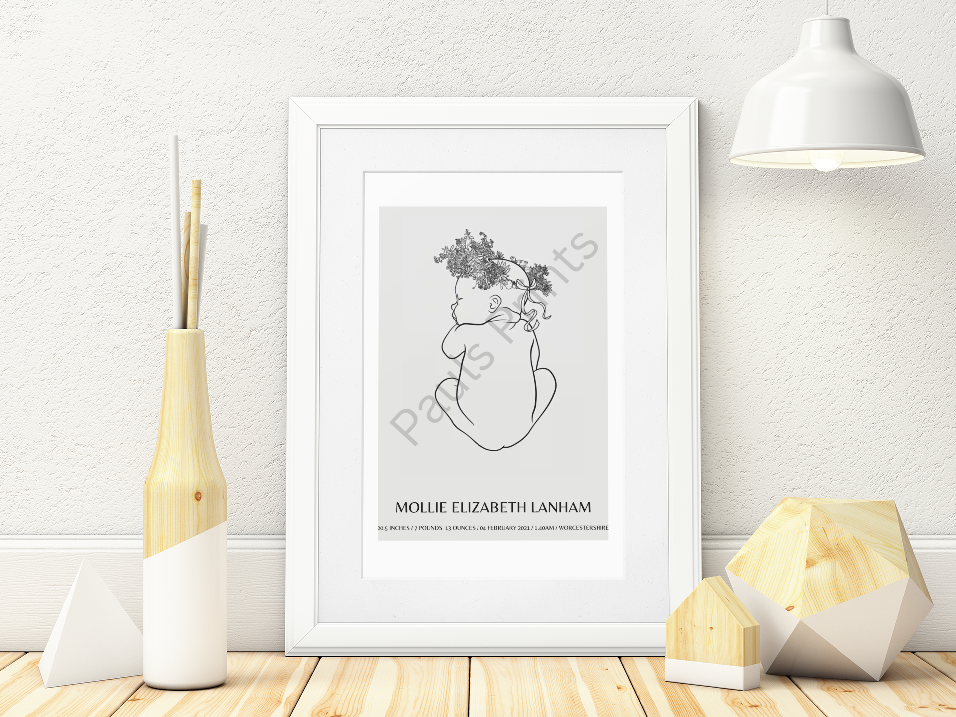Custom floral-crowned baby art print in a white frame, styled beside minimalist wooden ornaments and a pendant lamp.