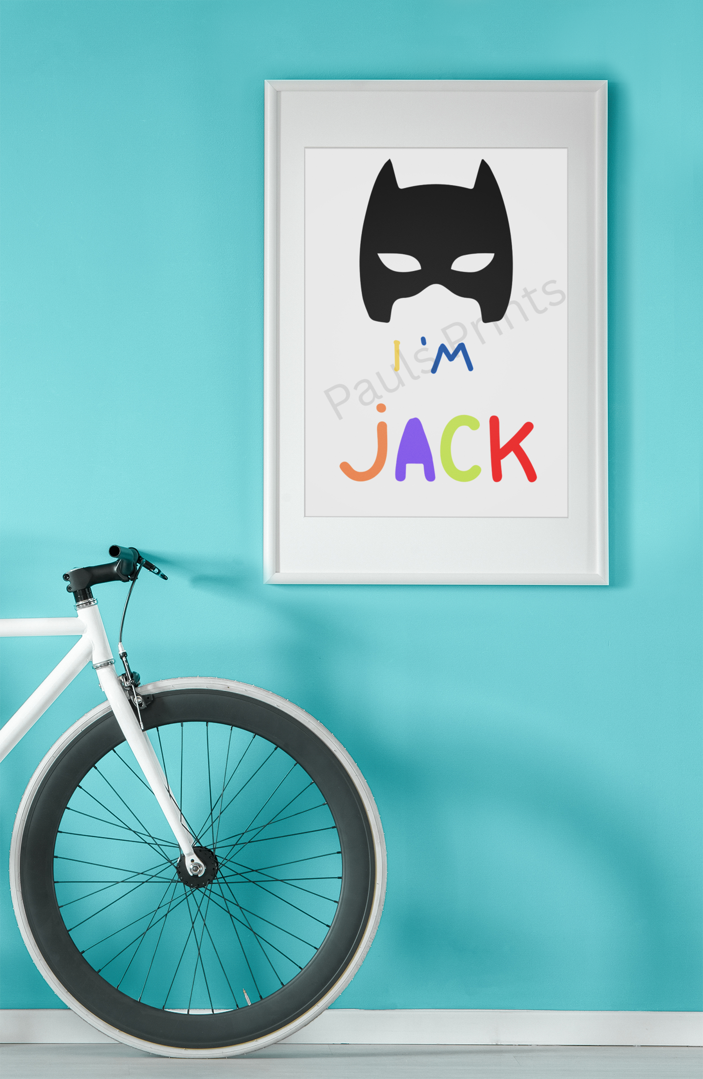 White-framed Batman mask print with “I’m Jack” in bold colours, displayed against a turquoise wall beside a white bicycle.
