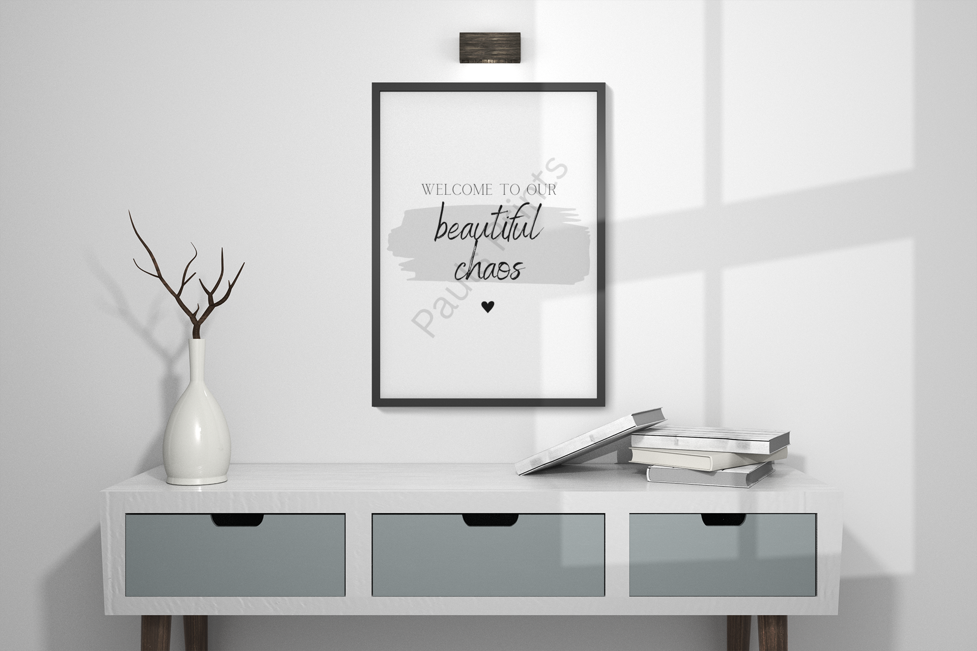 Monochrome “Welcome to our beautiful chaos” print styled above light wood console with books and ceramic vase.