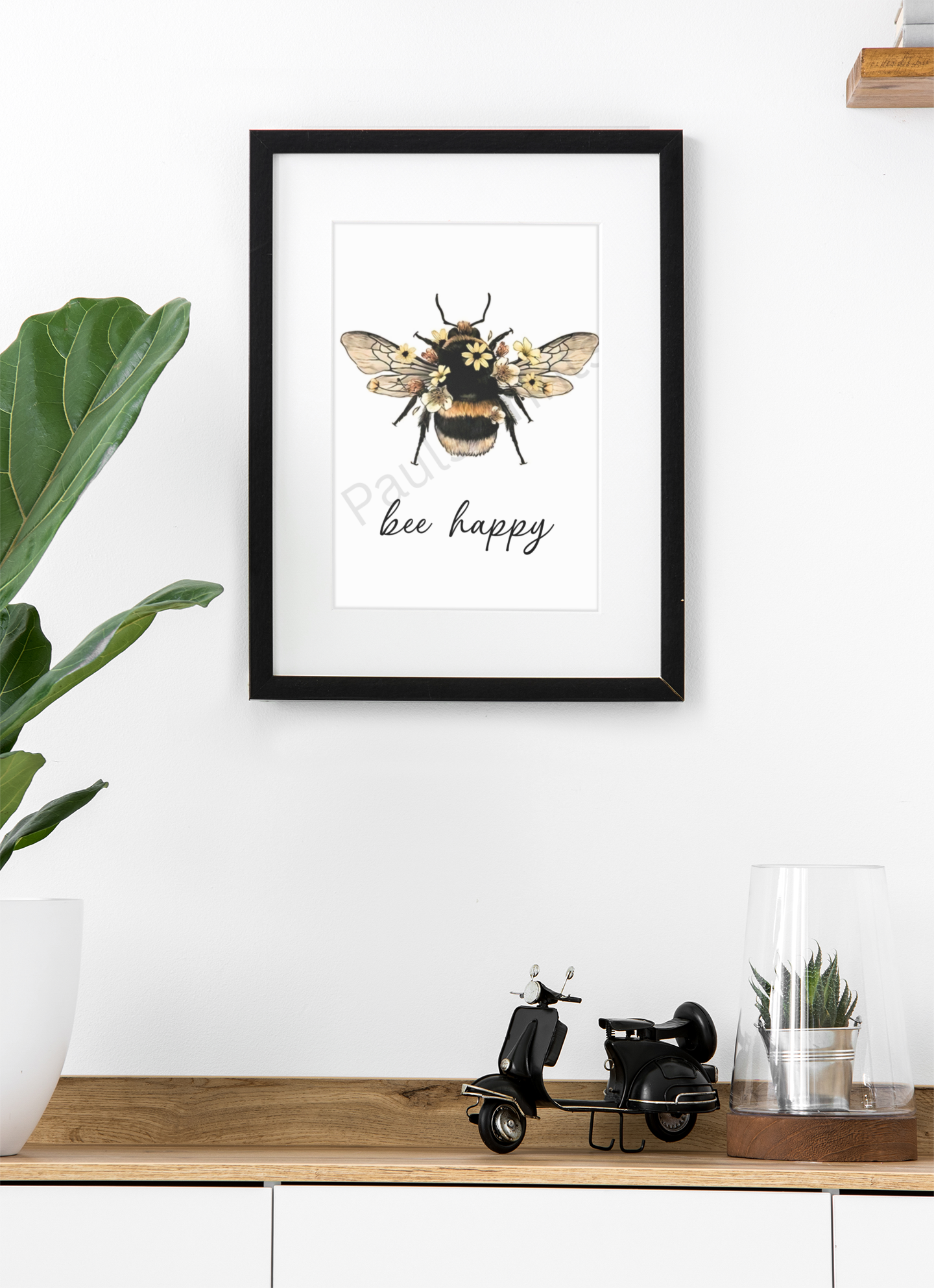 Framed “bee happy” art print with bee and flowers, hanging above a wooden cabinet with potted plant and miniature black scooter.