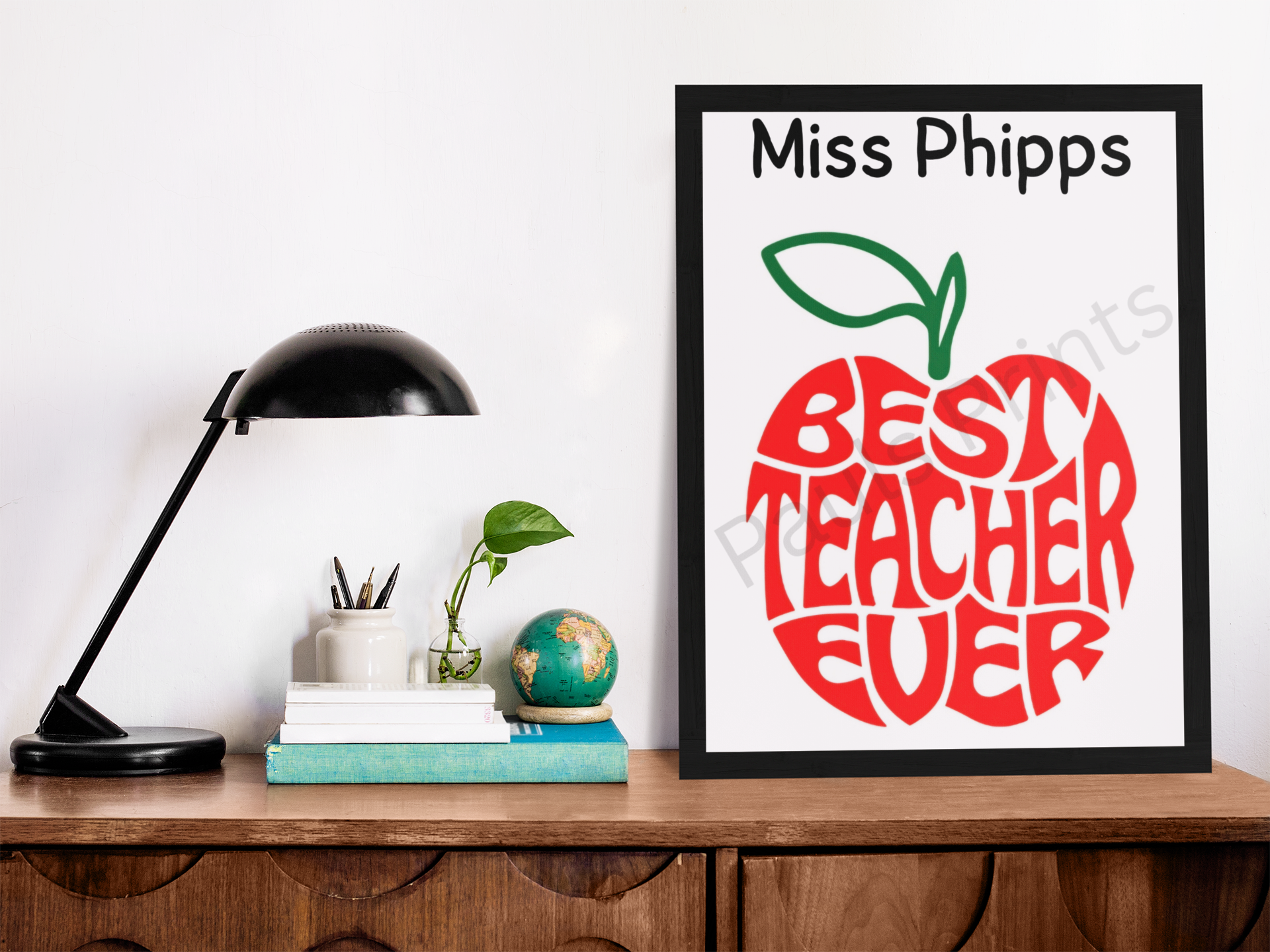Large black-framed “Best Teacher Ever” red apple print with teacher’s name, displayed on a wooden desk with lamp, books, and globe.