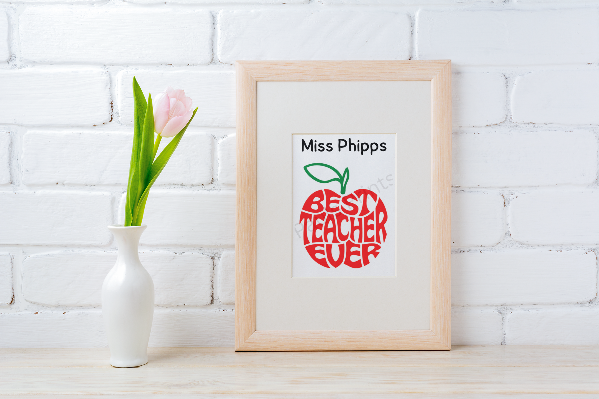 Light wood frame with “Best Teacher Ever” red apple print and teacher’s name, placed beside a white vase with pink tulip on white brick background.