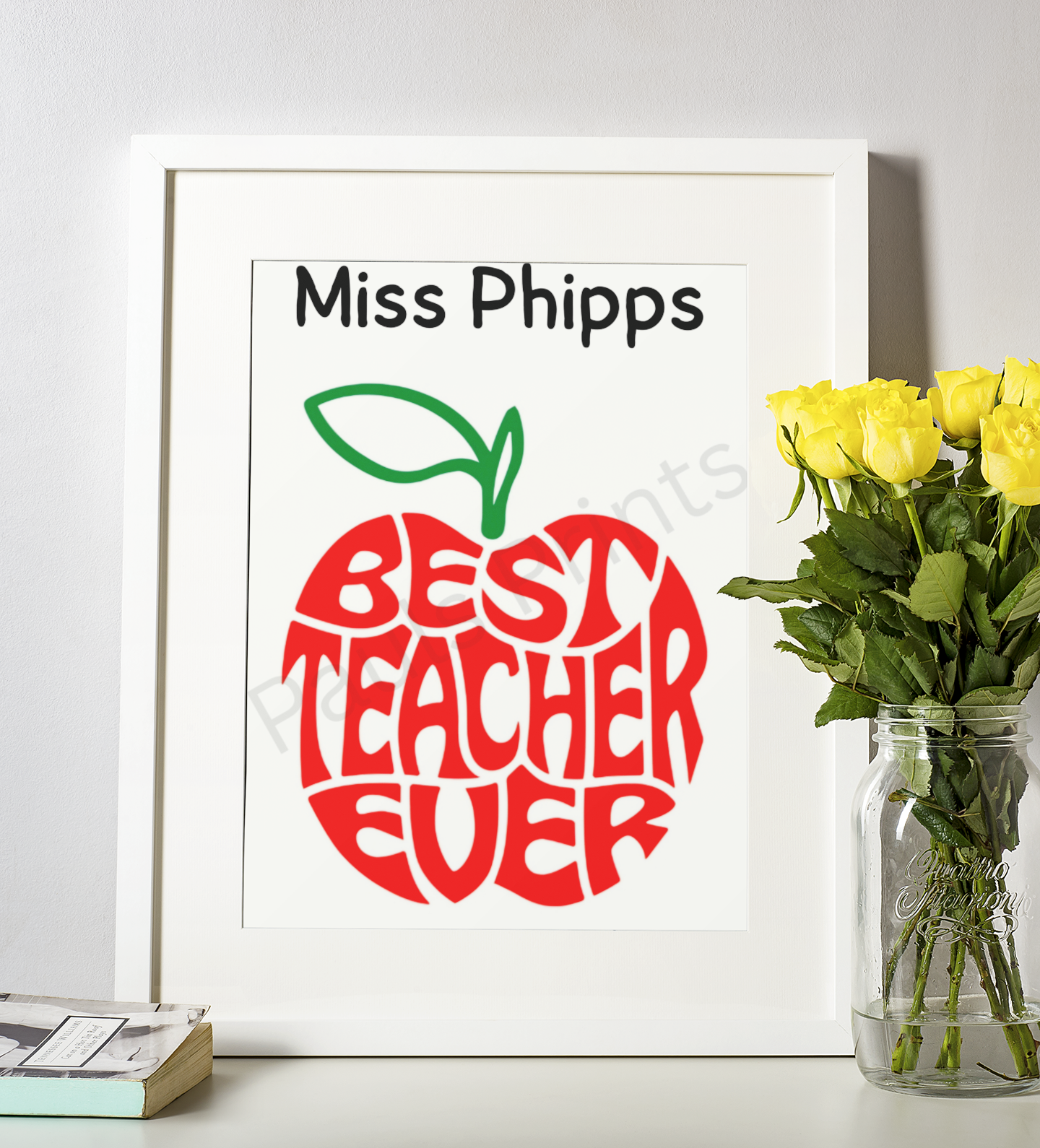 White-framed “Best Teacher Ever” apple print with teacher’s name, displayed beside a jar of yellow roses on a desk.
