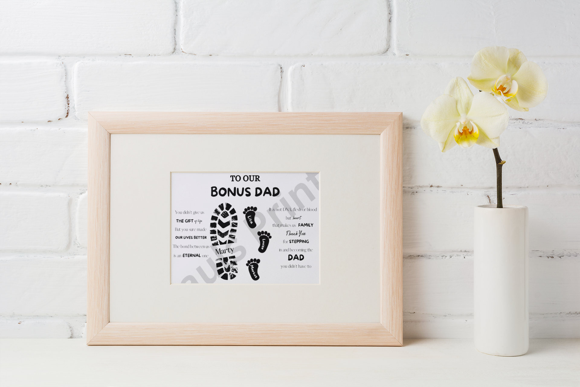 Small “To Our Bonus Dad” framed print with black footprint artwork placed on a white shelf next to a cream orchid in a modern minimalist setting.