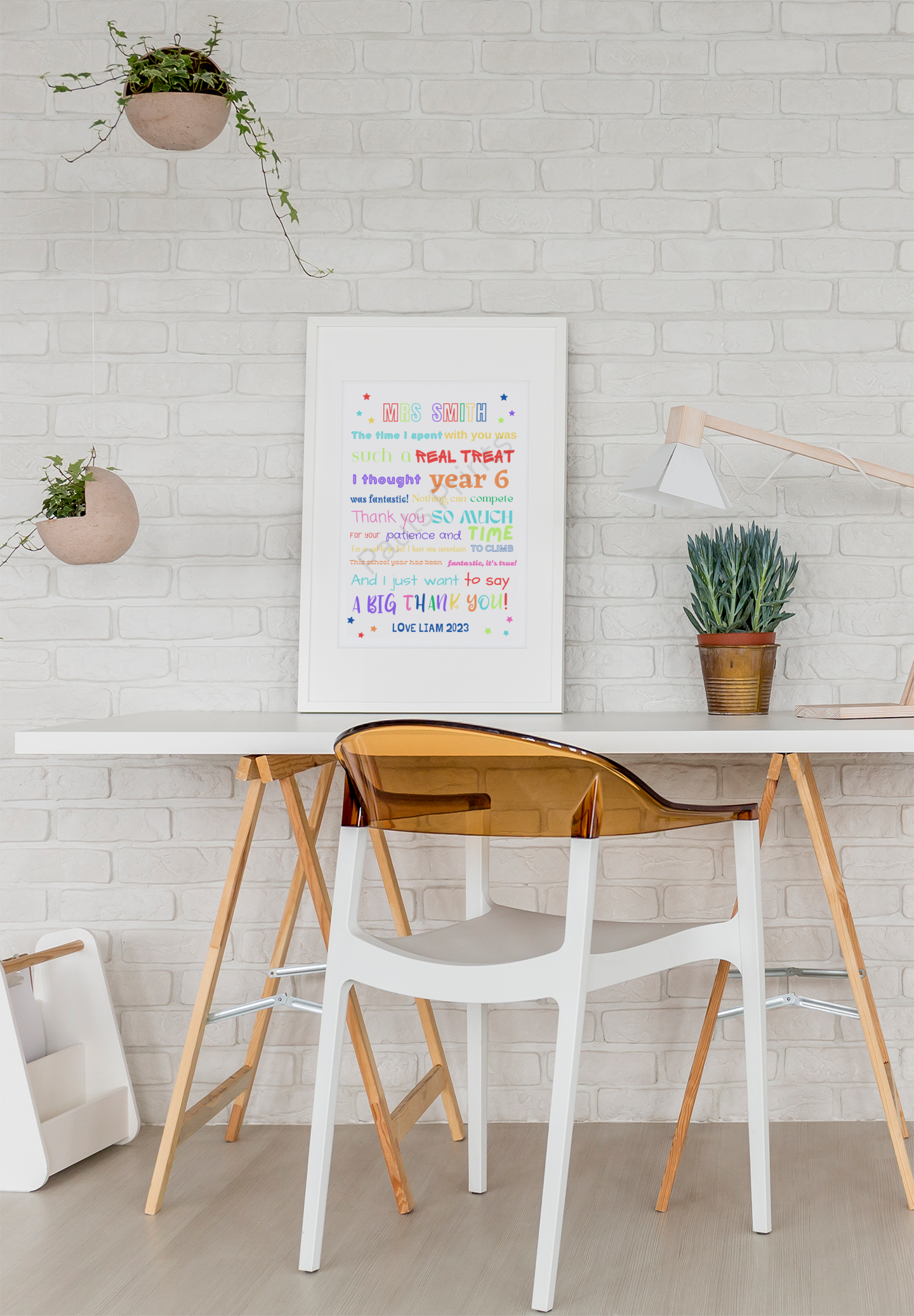 Framed multicoloured teacher thank you print on a modern desk setup with white brick wall, plants and wood elements.
