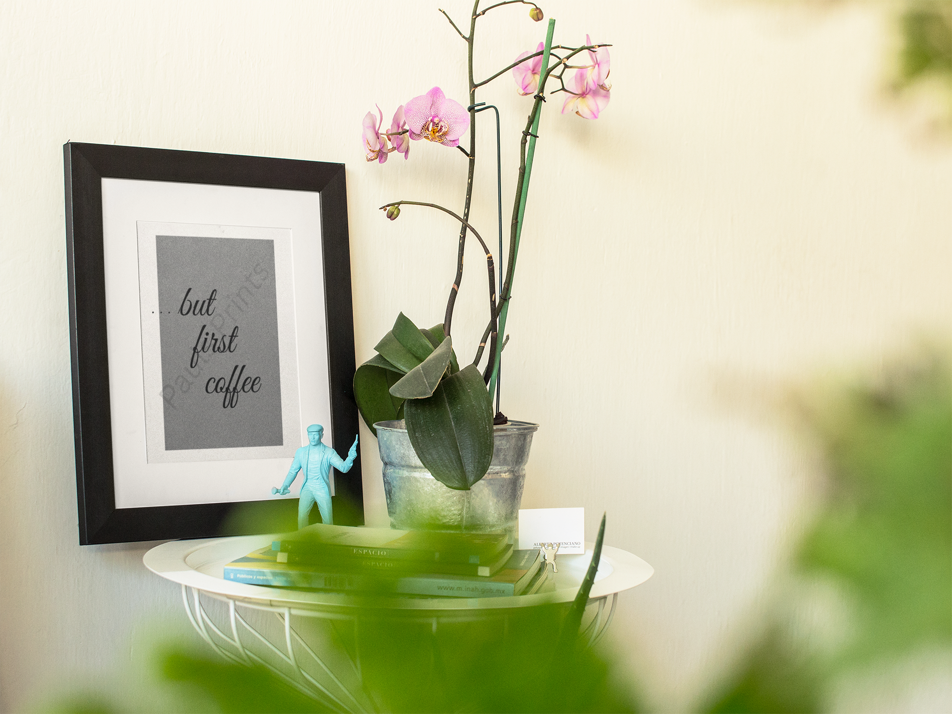 Black framed coffee quote print styled beside a potted orchid and books on a white metal side table.