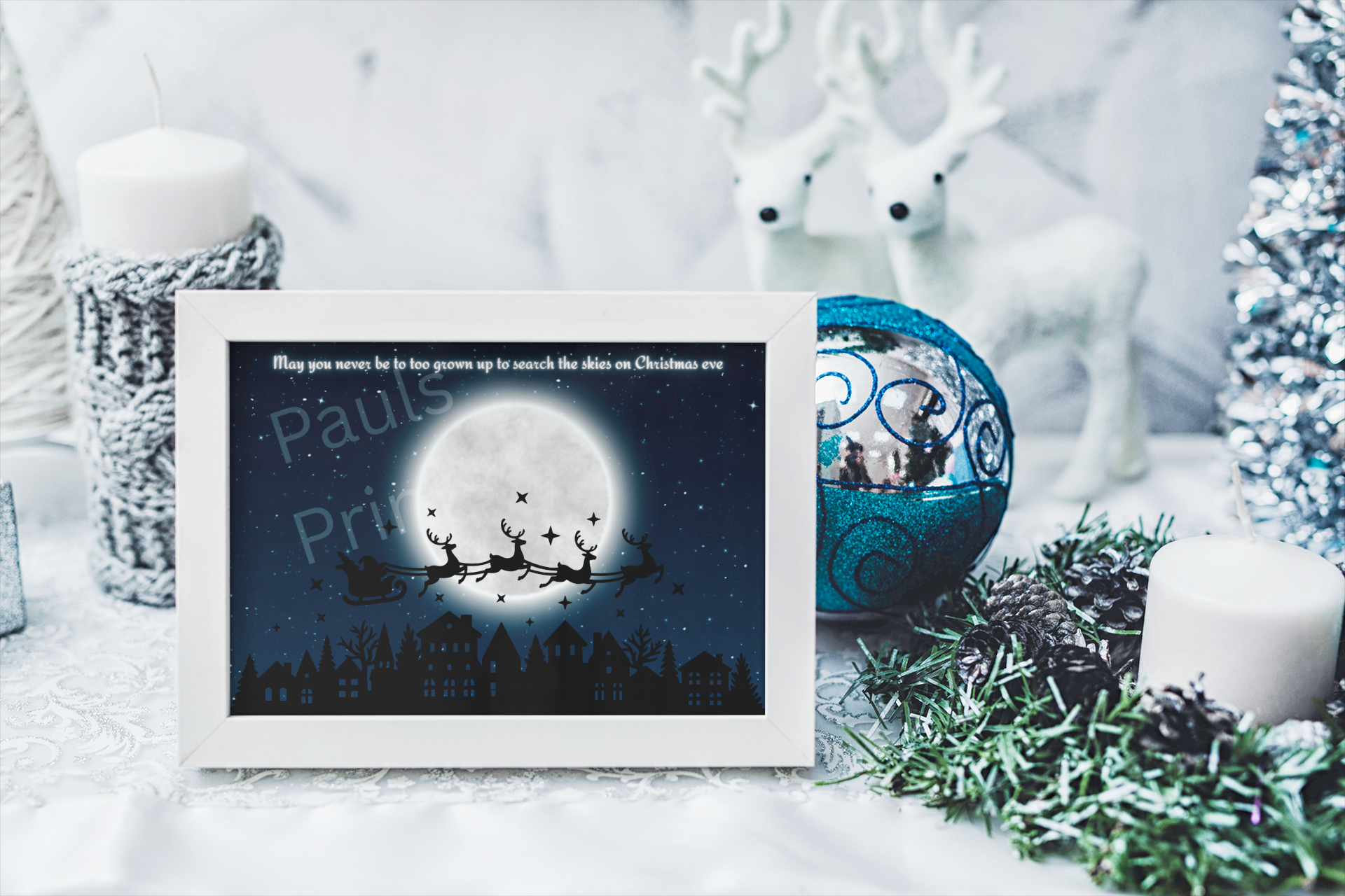 White-framed Christmas Eve Santa sleigh print on a snowy table with candles, reindeer ornaments, pinecones, and a blue Christmas bauble.