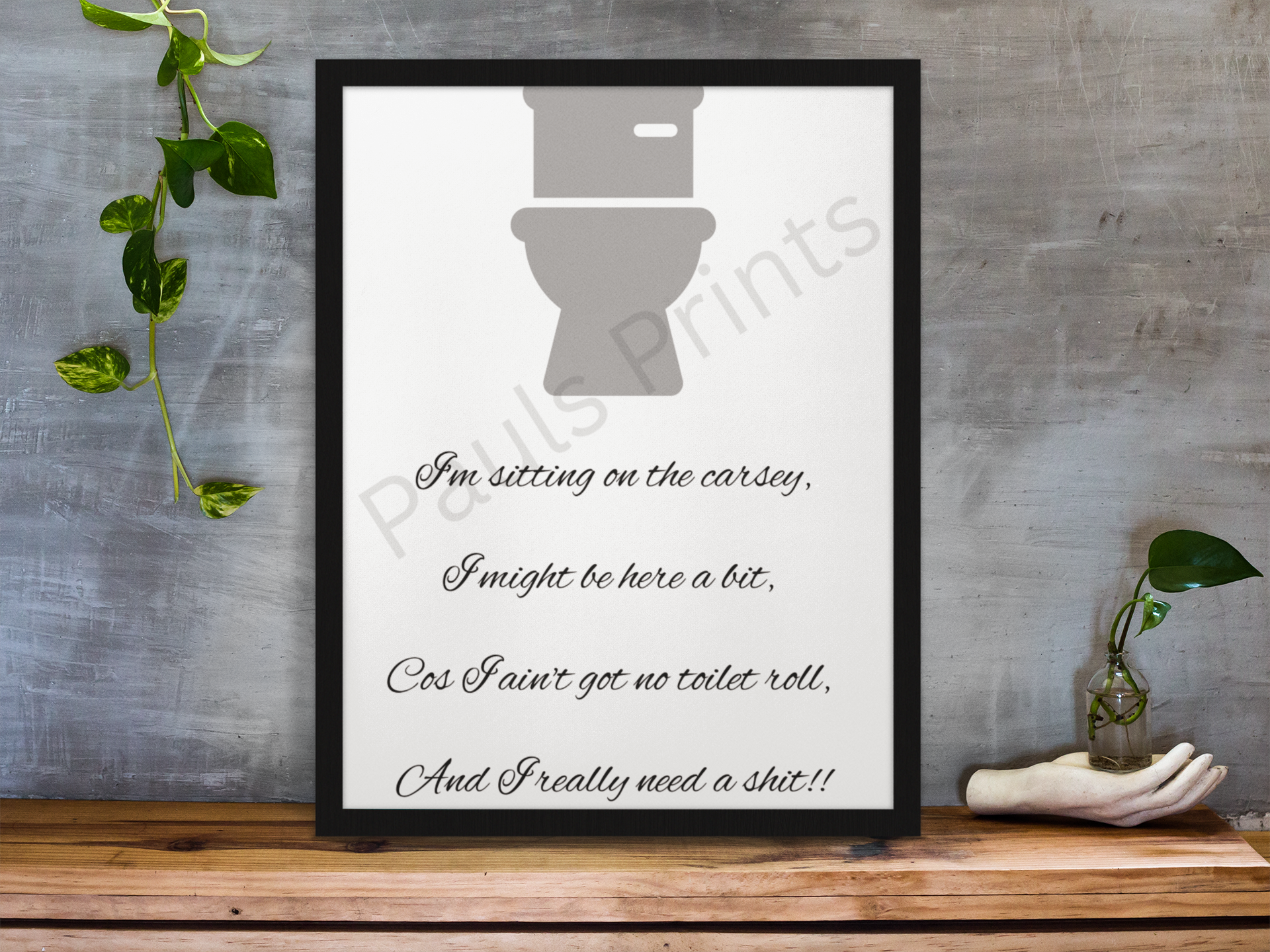 Framed funny bathroom quote print with grey toilet graphic, styled on a rustic wooden shelf with plants and grey wall backdrop.