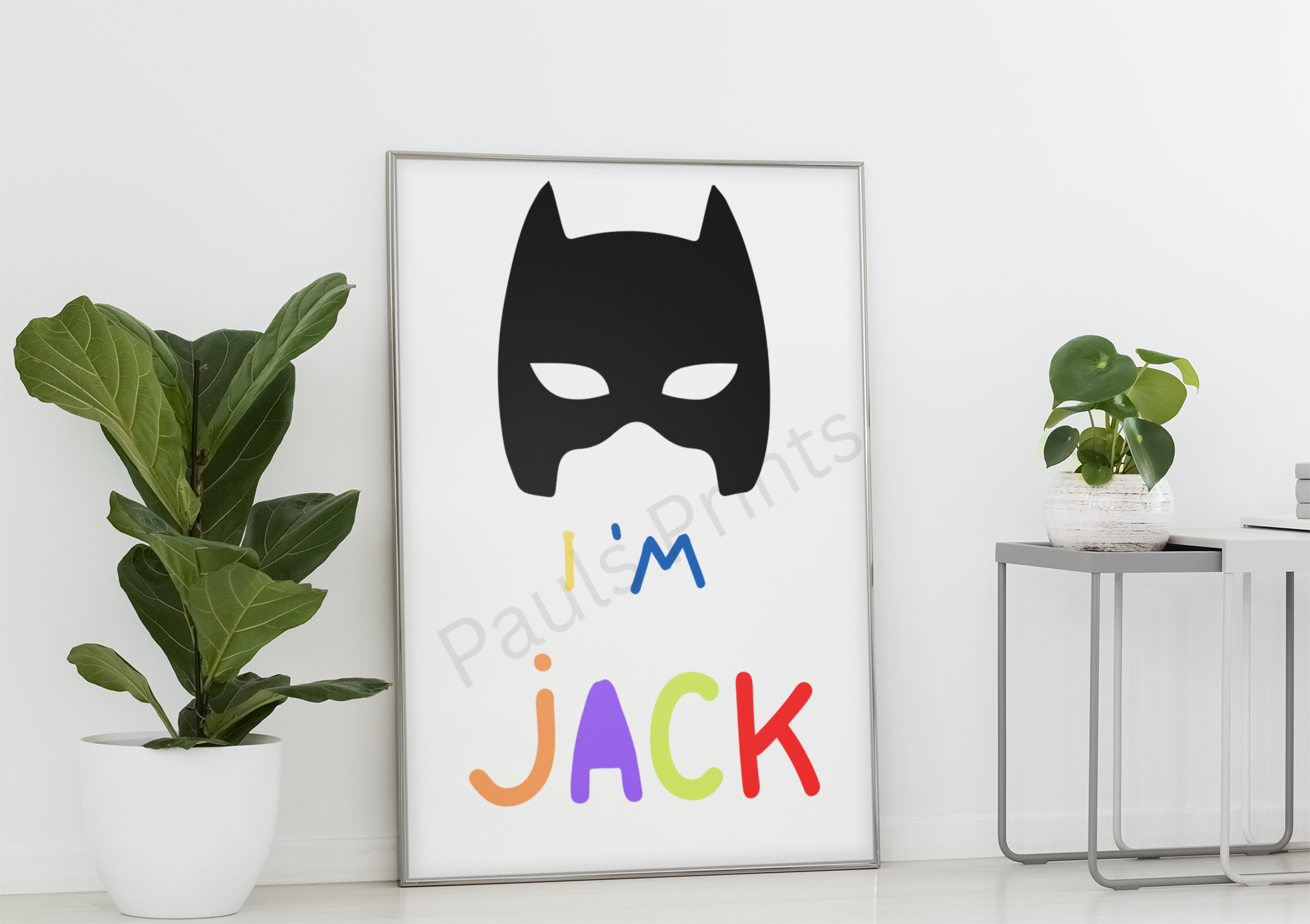 Minimalist Batman mask wall print with “I’m Jack” in playful colours, leaning against a white wall beside a potted plant.