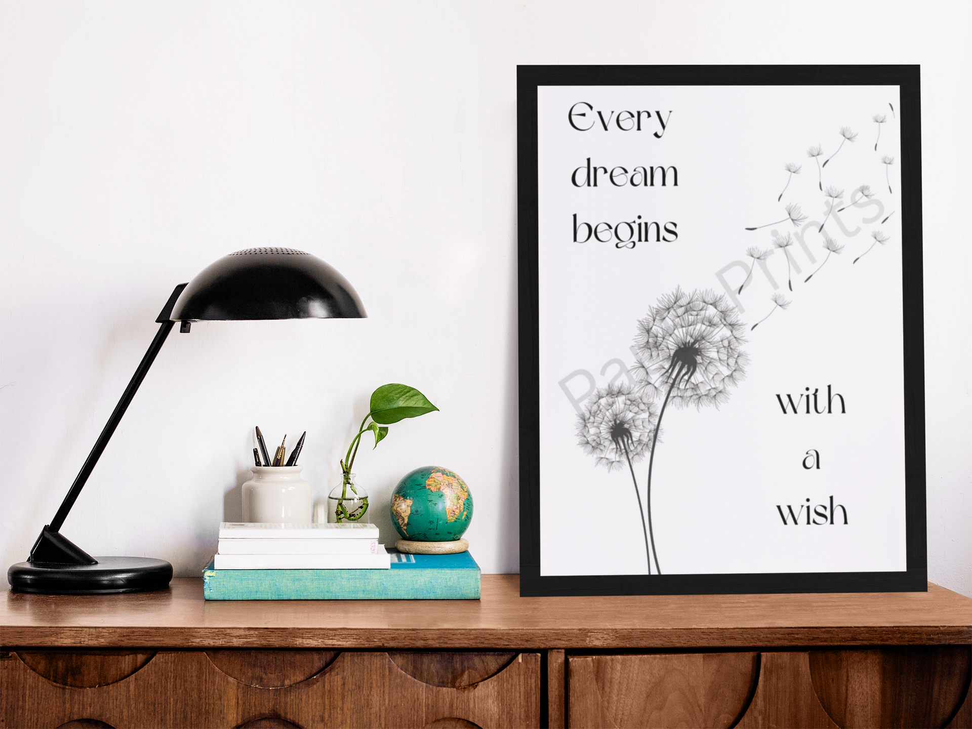 Black-framed dandelion quote print on a wooden desk with a black lamp, books, globe, and potted plant.