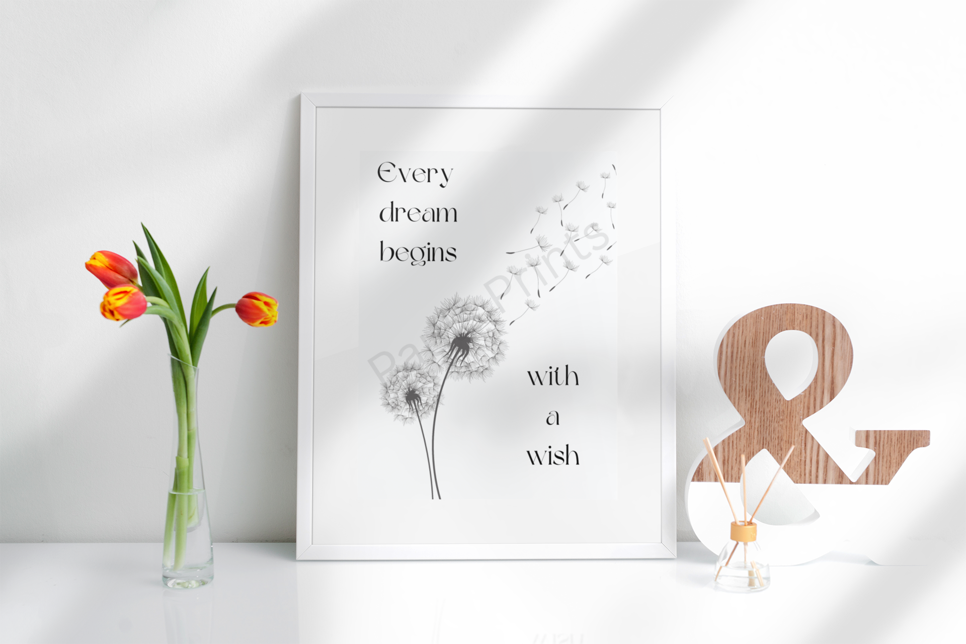 Framed dandelion print with "Every dream begins with a wish" quote, styled on a white surface beside tulips and a wooden ampersand.
