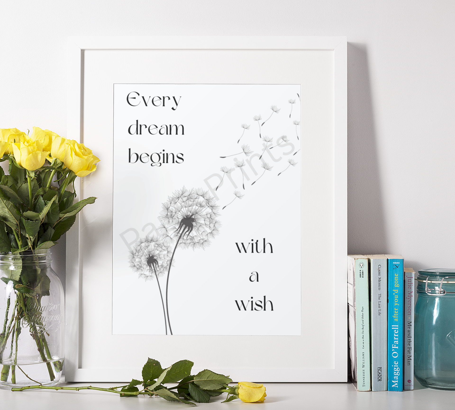 Framed inspirational dandelion print beside a bunch of yellow roses and books on a white surface.