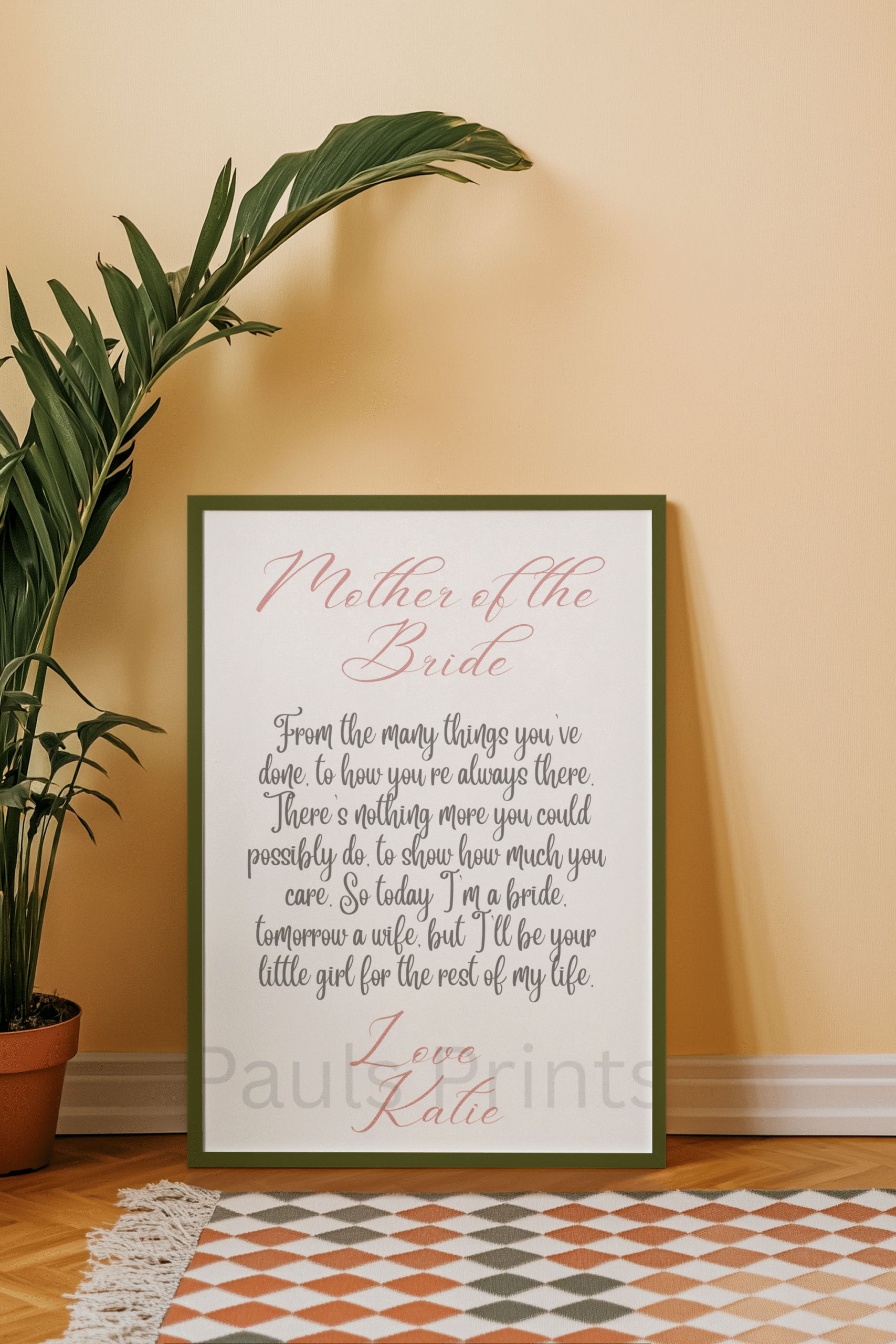 Mother of the Bride poem print in a deep green frame leaning against a peach-colored wall with leafy plants and a patterned rug.