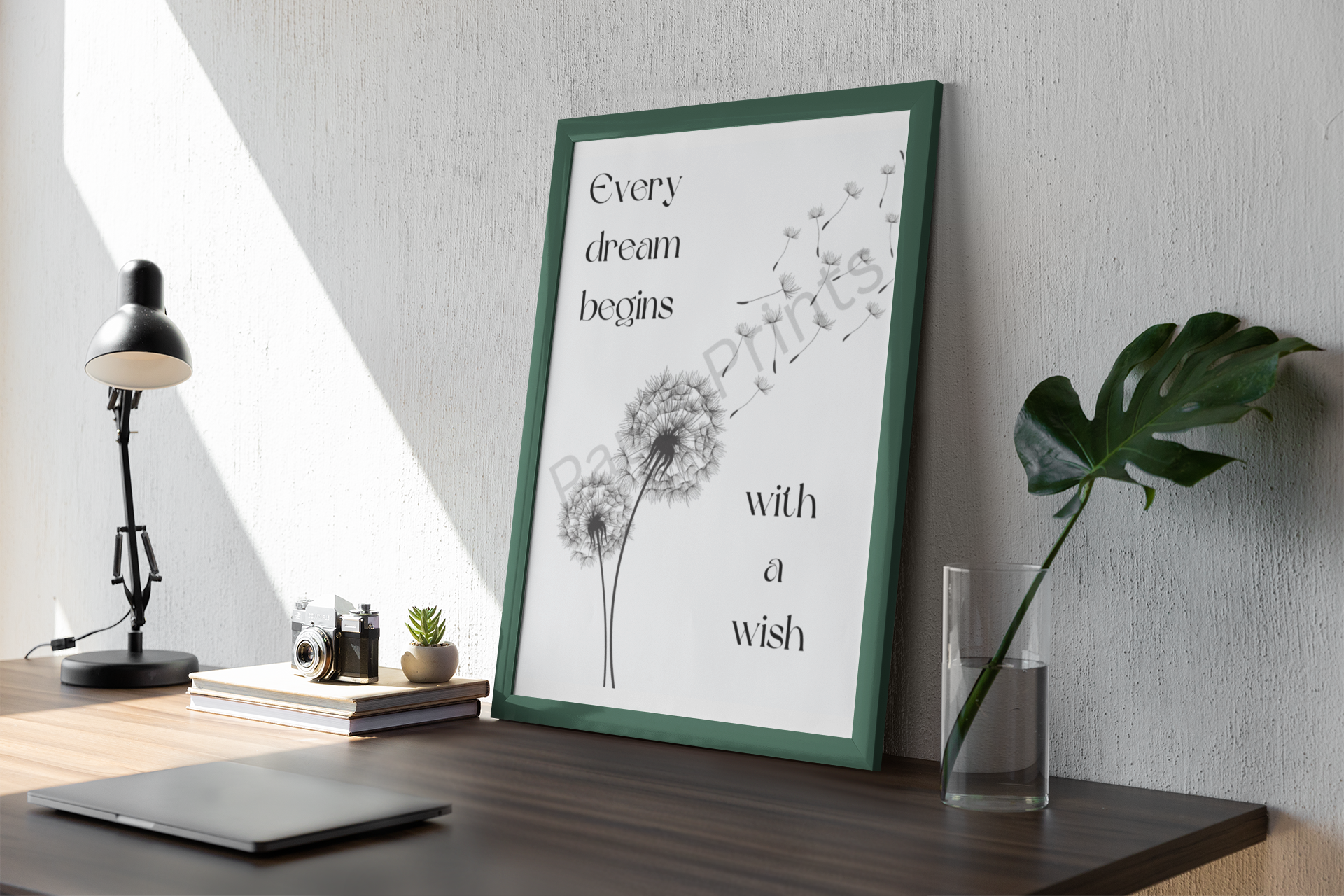 Framed dandelion quote print with green frame, placed on a modern desk beside a glass vase and a black lamp.