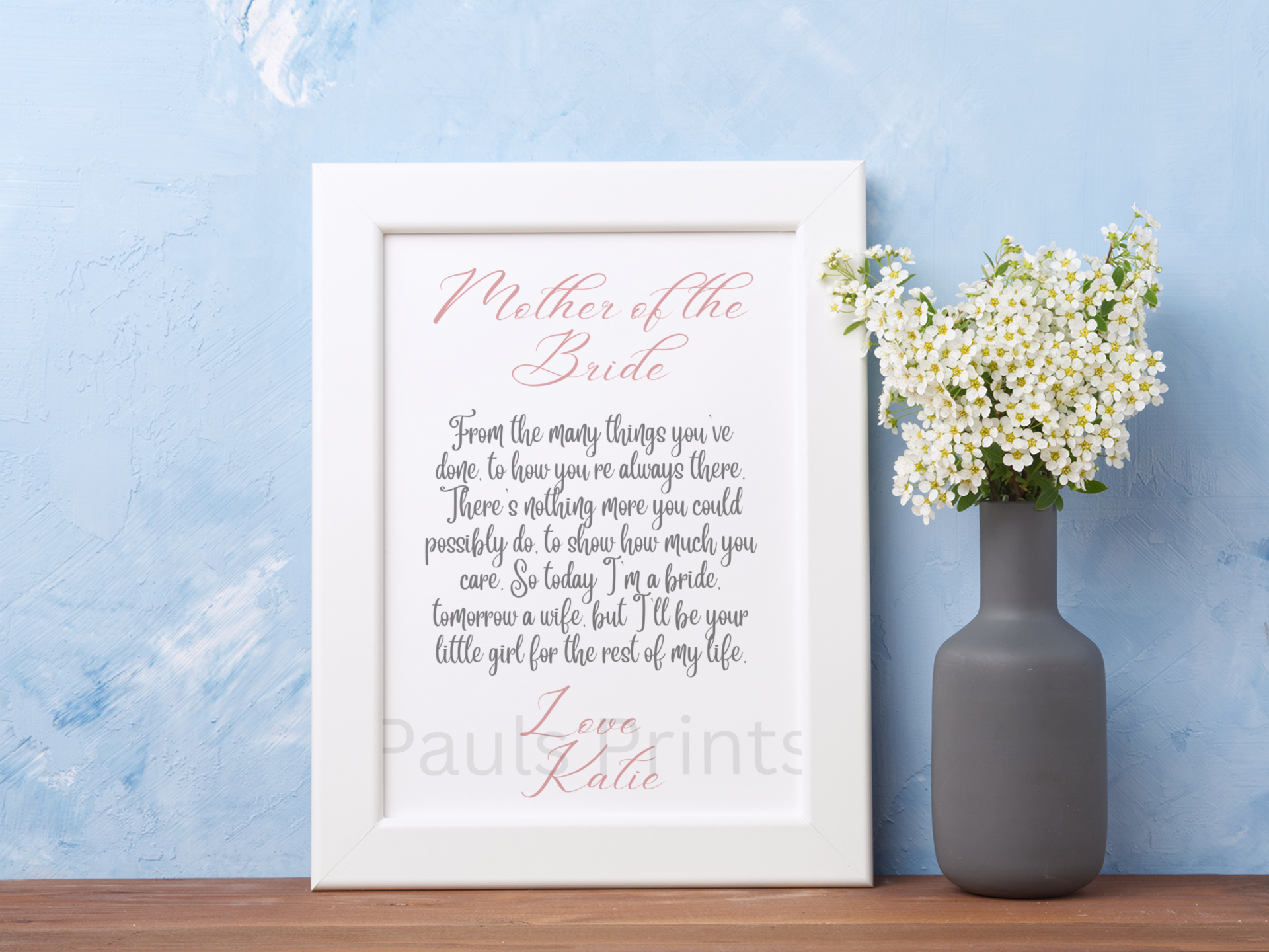 Wedding poem print in a white frame styled with a vase of white flowers on a wooden table against a soft blue background.