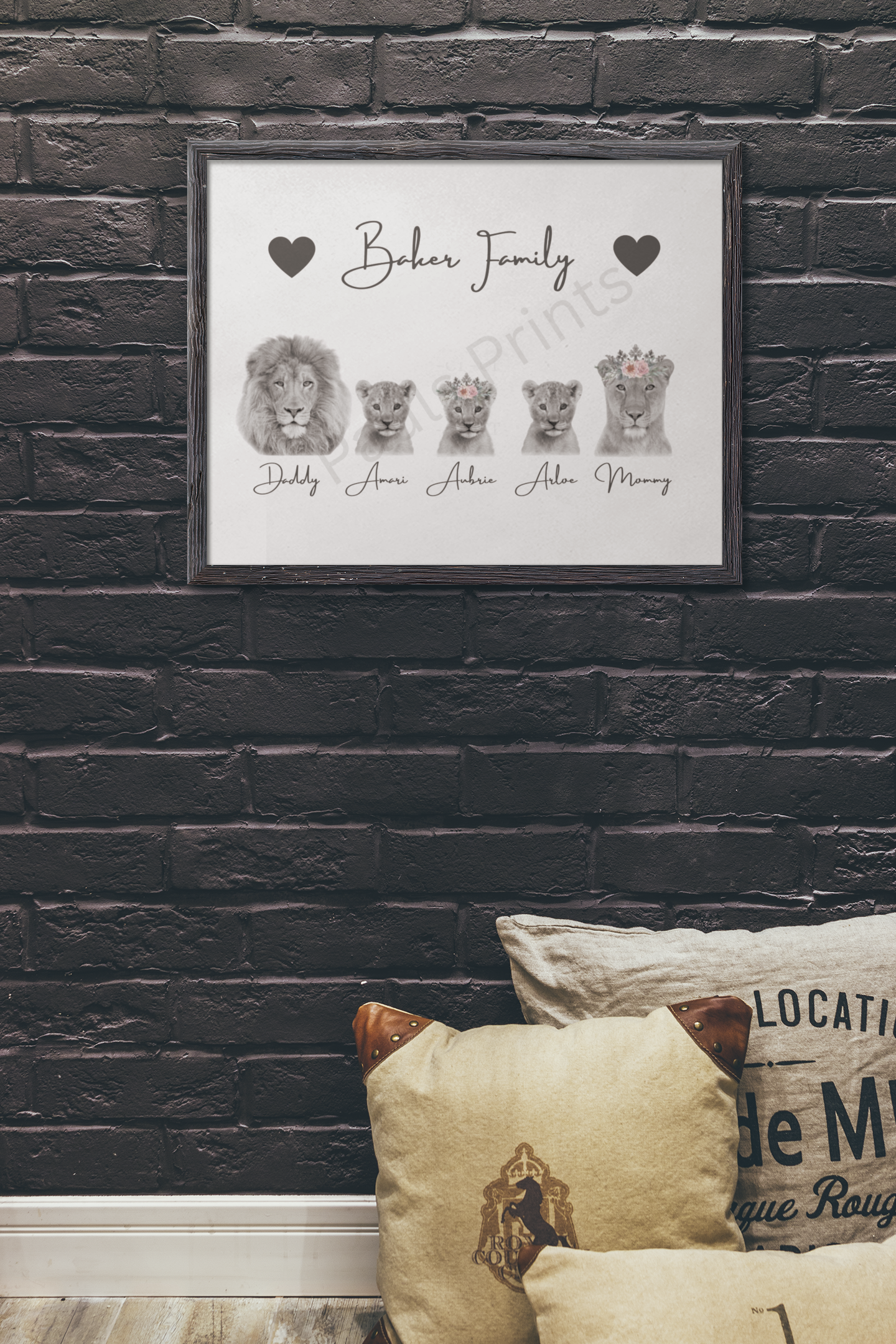 Framed lion family artwork with personalised names and heart designs, mounted on a dark brick wall above cushions.