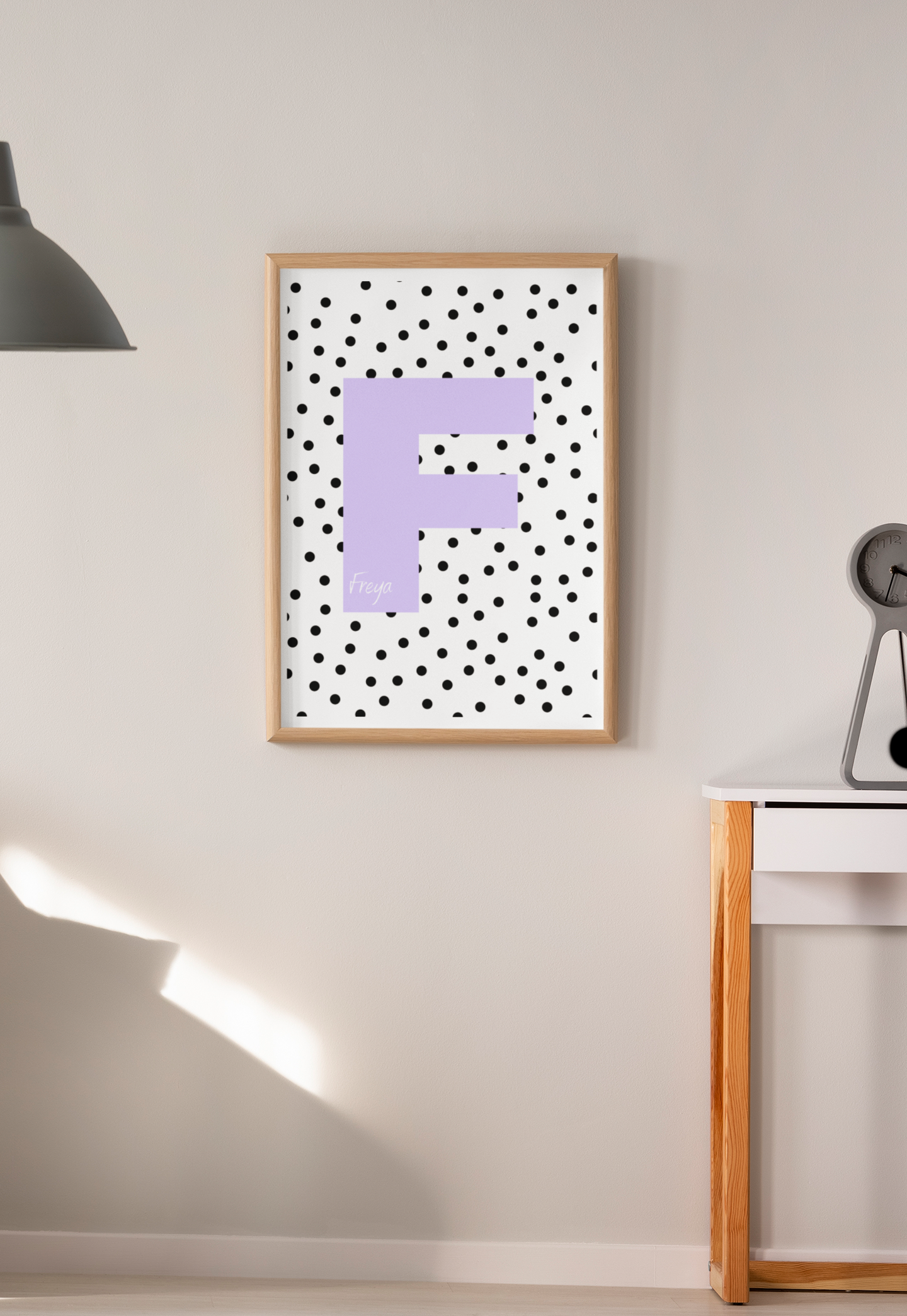 Lavender letter F wall print with black polka dots, displayed in wooden frame on beige wall near modern desk and lamp.