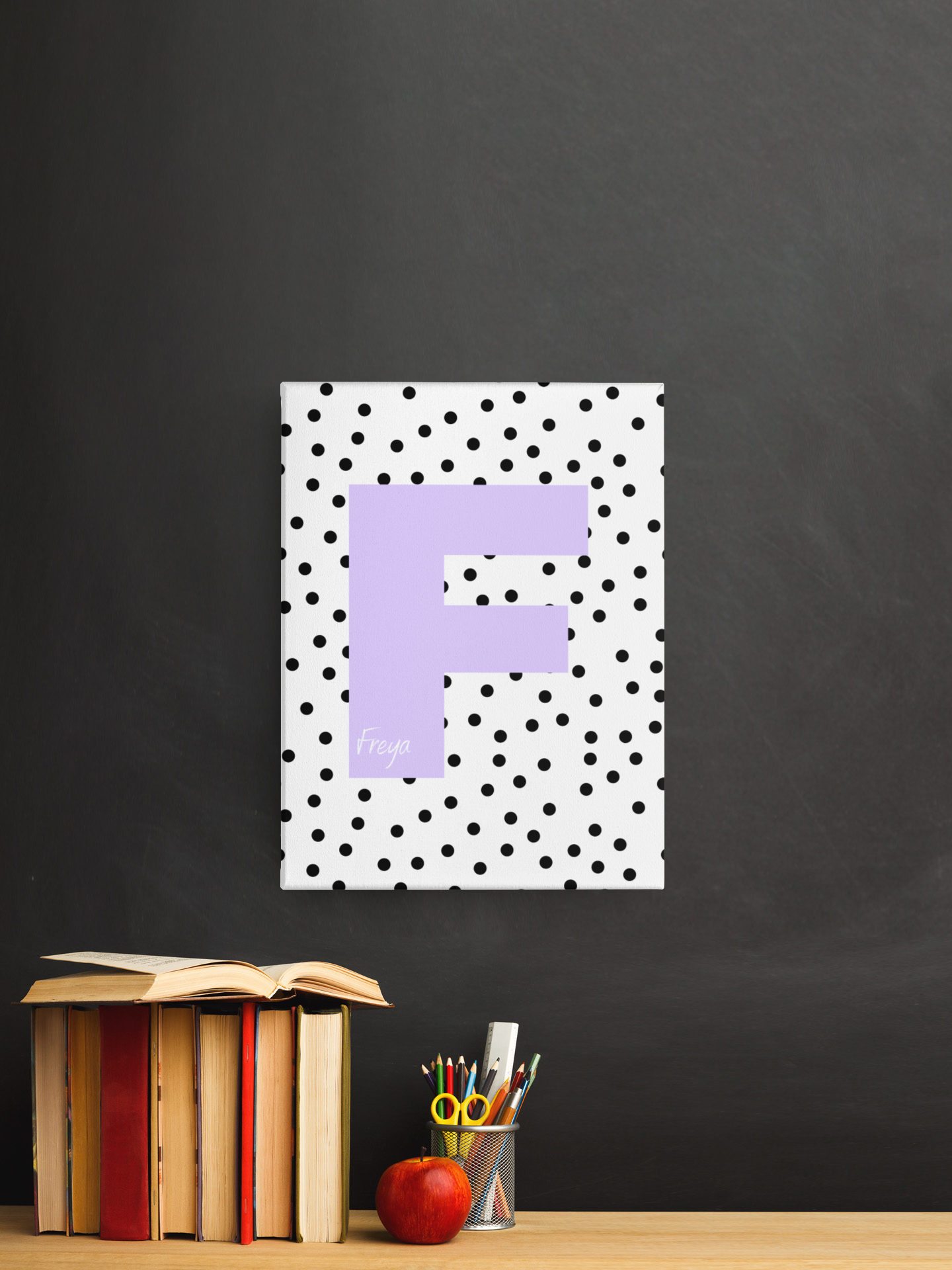 Framed letter F wall art with black polka dots on white background, featuring name Freya in lavender, displayed on blackboard wall above desk with books and apple.
