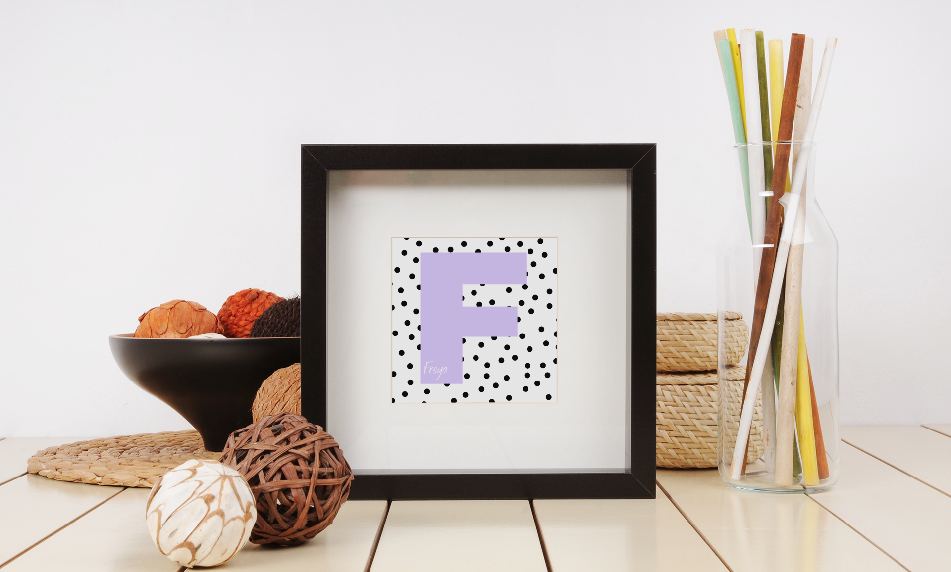Black-framed letter F print with lavender initial and black polka dot background, displayed beside wicker baskets, vase, and decorative balls.
