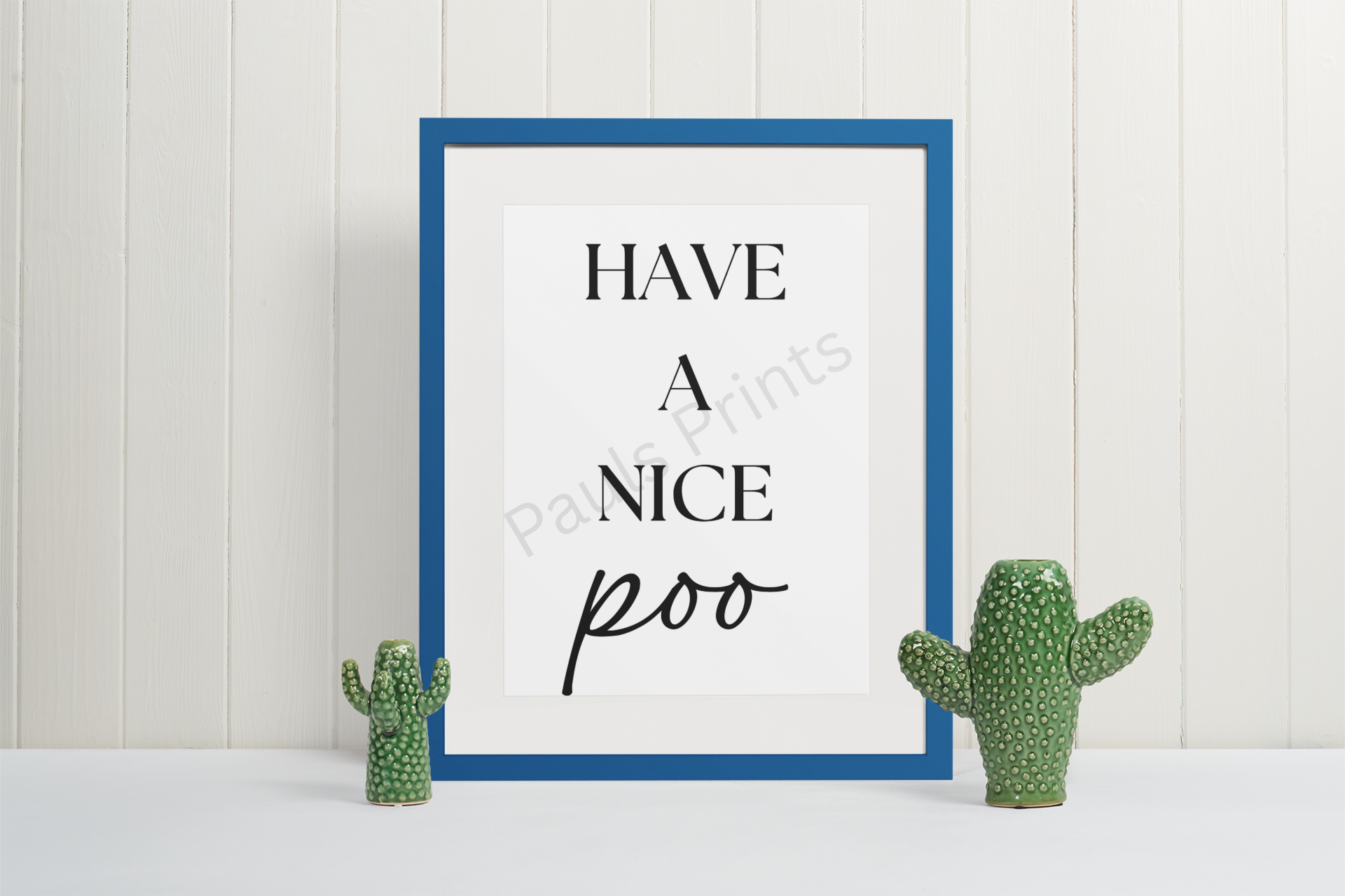 Minimalist bathroom art print with the text “Have a Nice Poo,” framed in blue, placed between two small cactus ornaments on a white background.