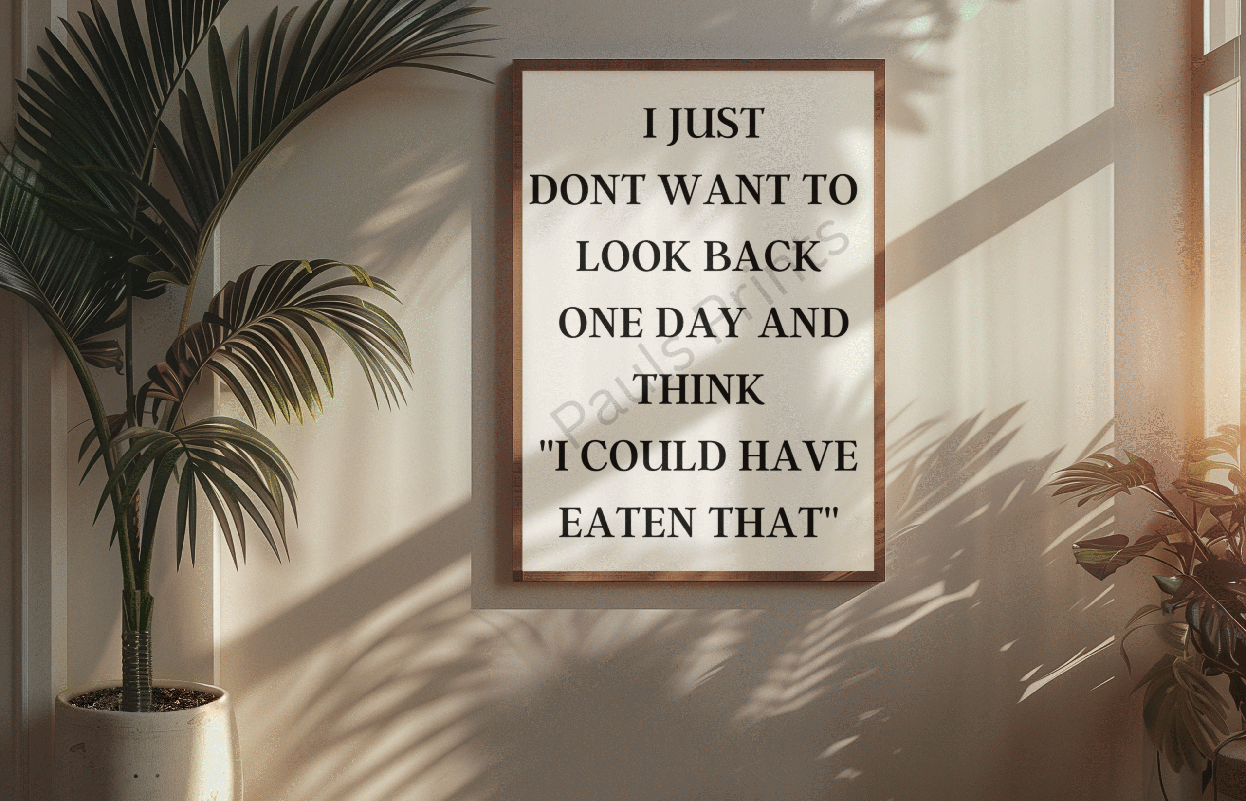 Framed quote print saying “I just don’t want to look back one day and think I could have eaten that” on a light wall with large tropical plants and soft sunlight.