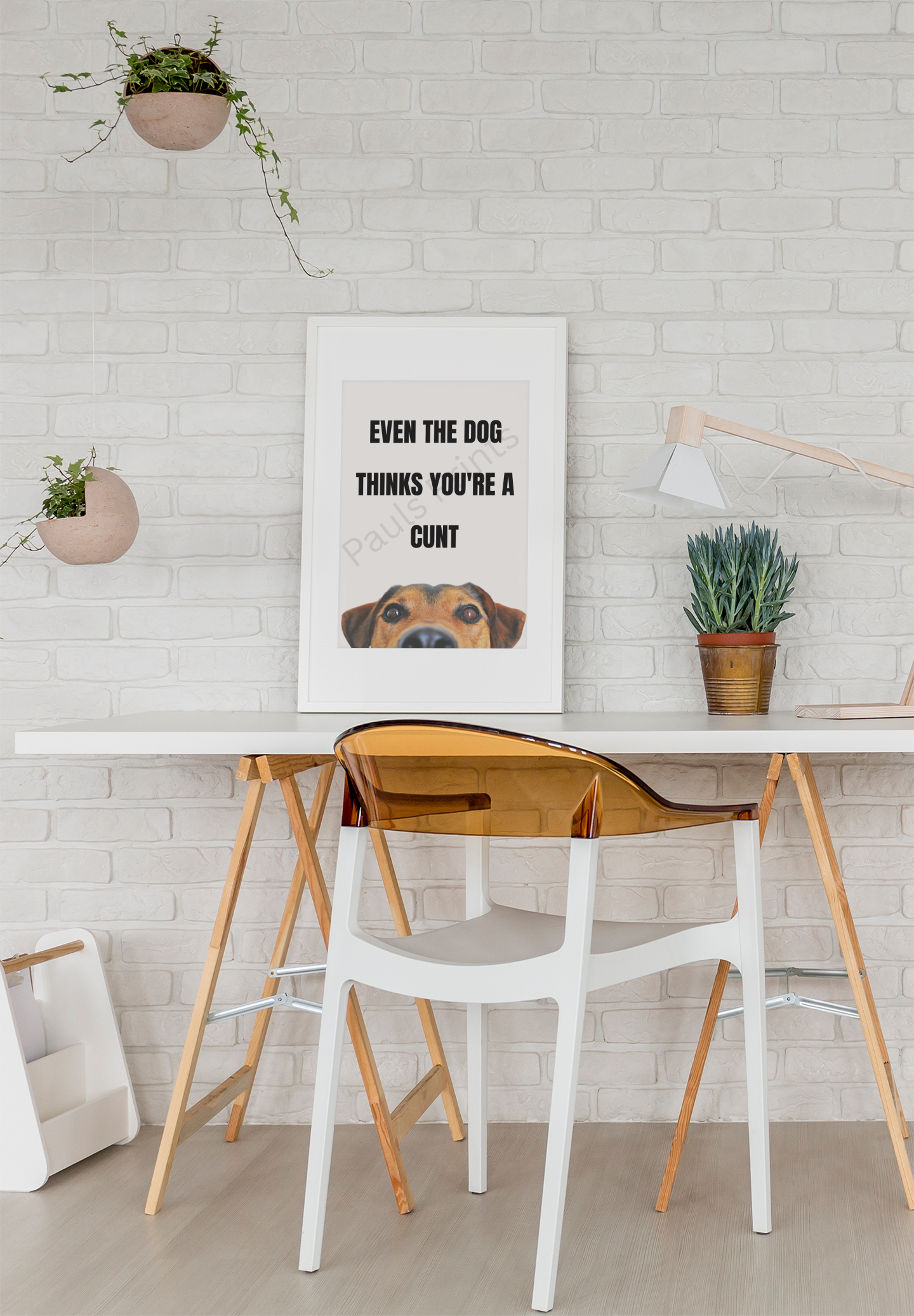 Humorous framed print with bold black text and a peeking dog illustration, displayed on a white brick wall above a modern desk with plants and accessories.