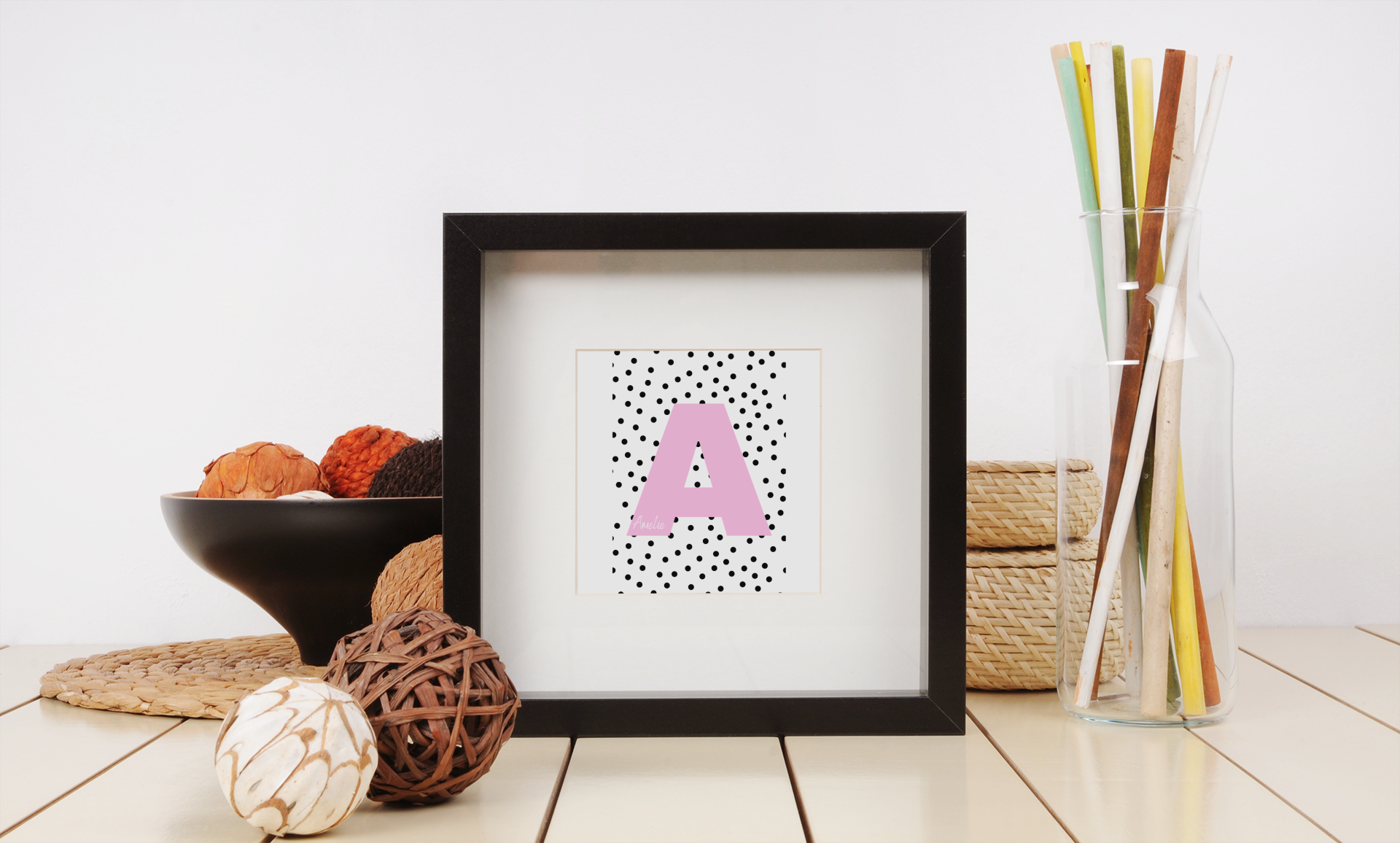 Framed wall print with pink A and playful dot background, styled with wooden flooring and indoor greenery.