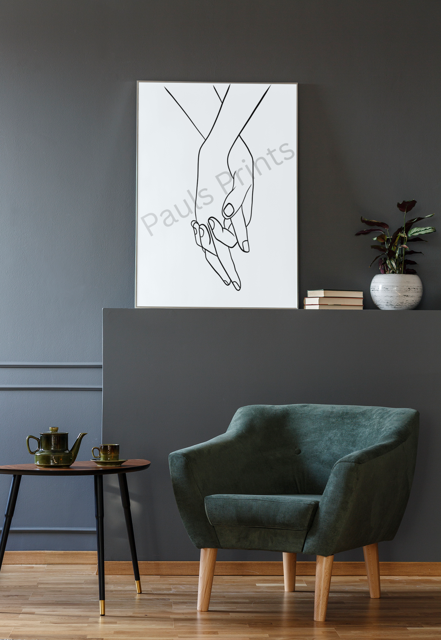 Black and white line drawing of two hands holding, framed and placed against a dark grey wall with a green armchair and small table.