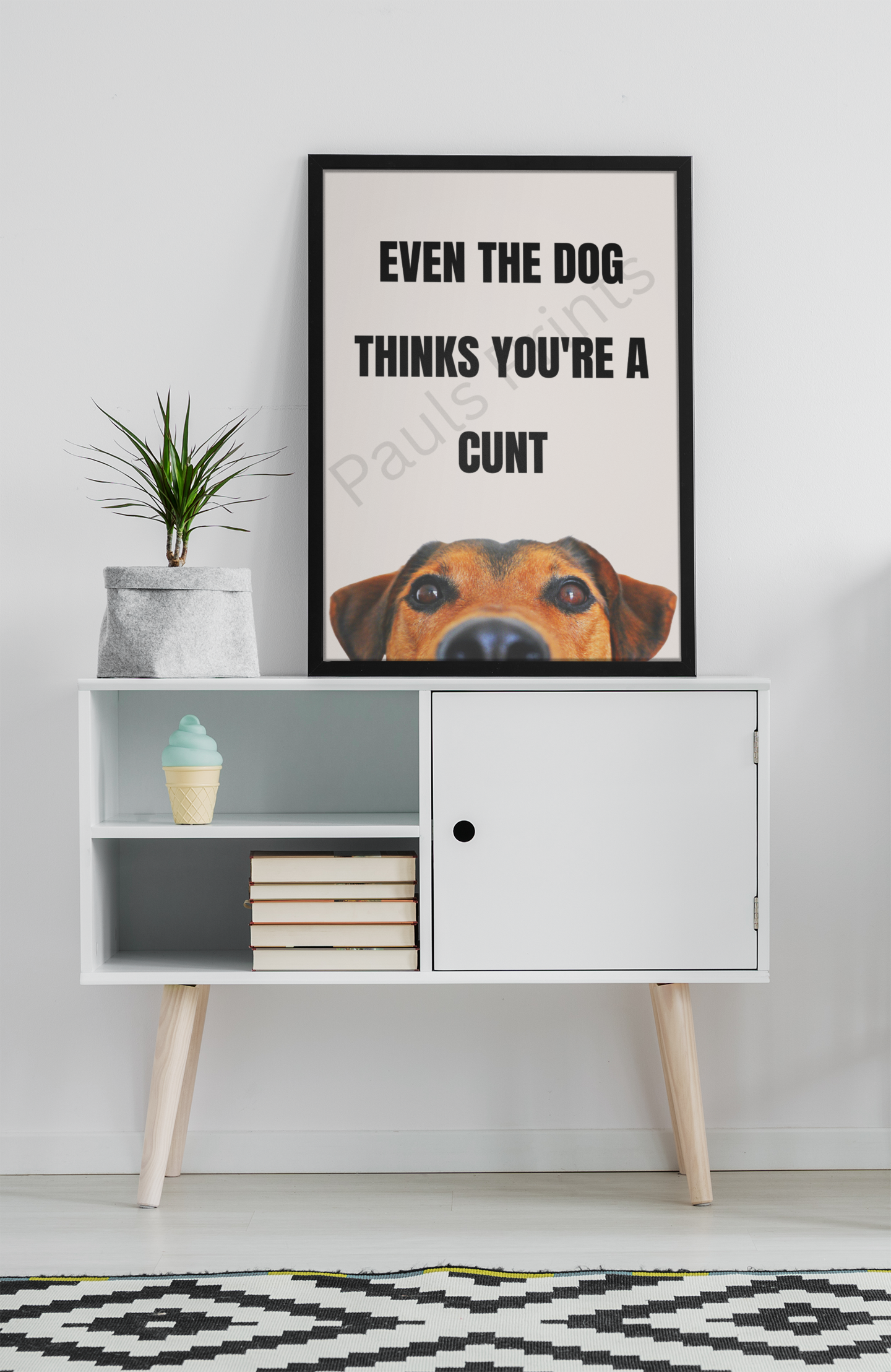Funny print saying “Even the dog thinks you’re a c***” with dog illustration, framed in black and placed on top of a modern white sideboard.